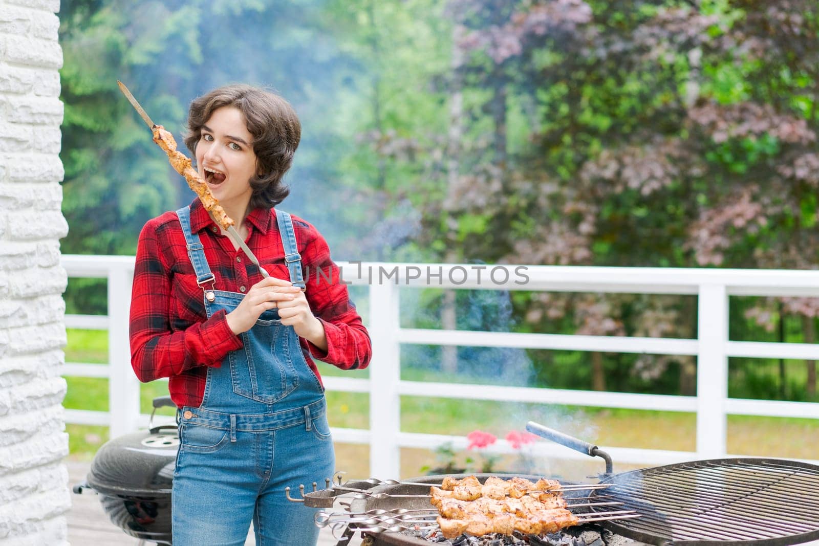 Barbeque party in garden with young woman in denim overalls and red plaid shirt in a country house on the terrace preparing meat skewers