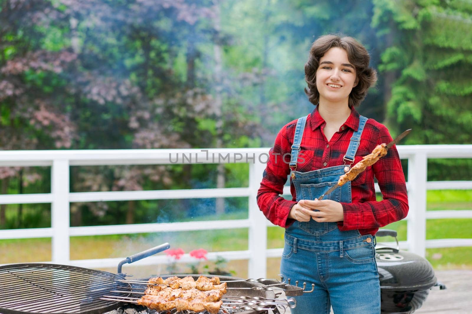 Barbeque party in garden with young woman in denim overalls and red plaid shirt in a country house on the terrace preparing meat skewers