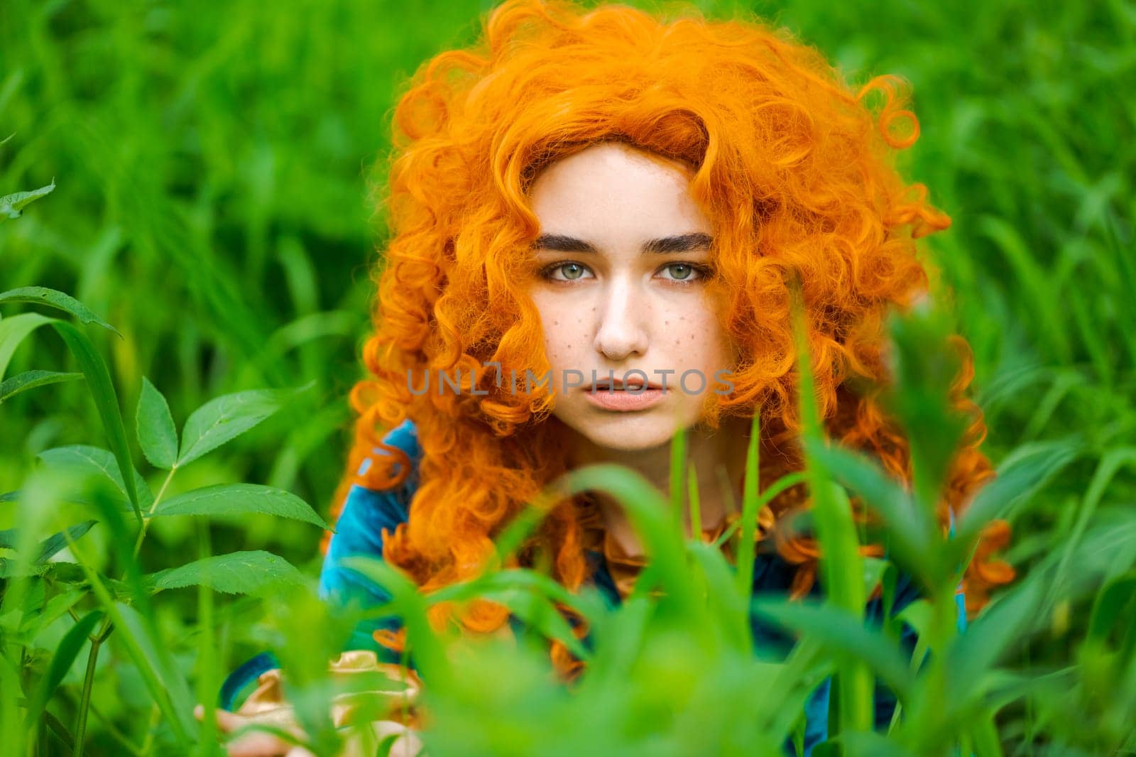 Cute girl in beautiful dress in an forest. Character cosplay festival portrait girl with curly long hair against the background of green bushes in a blue dress