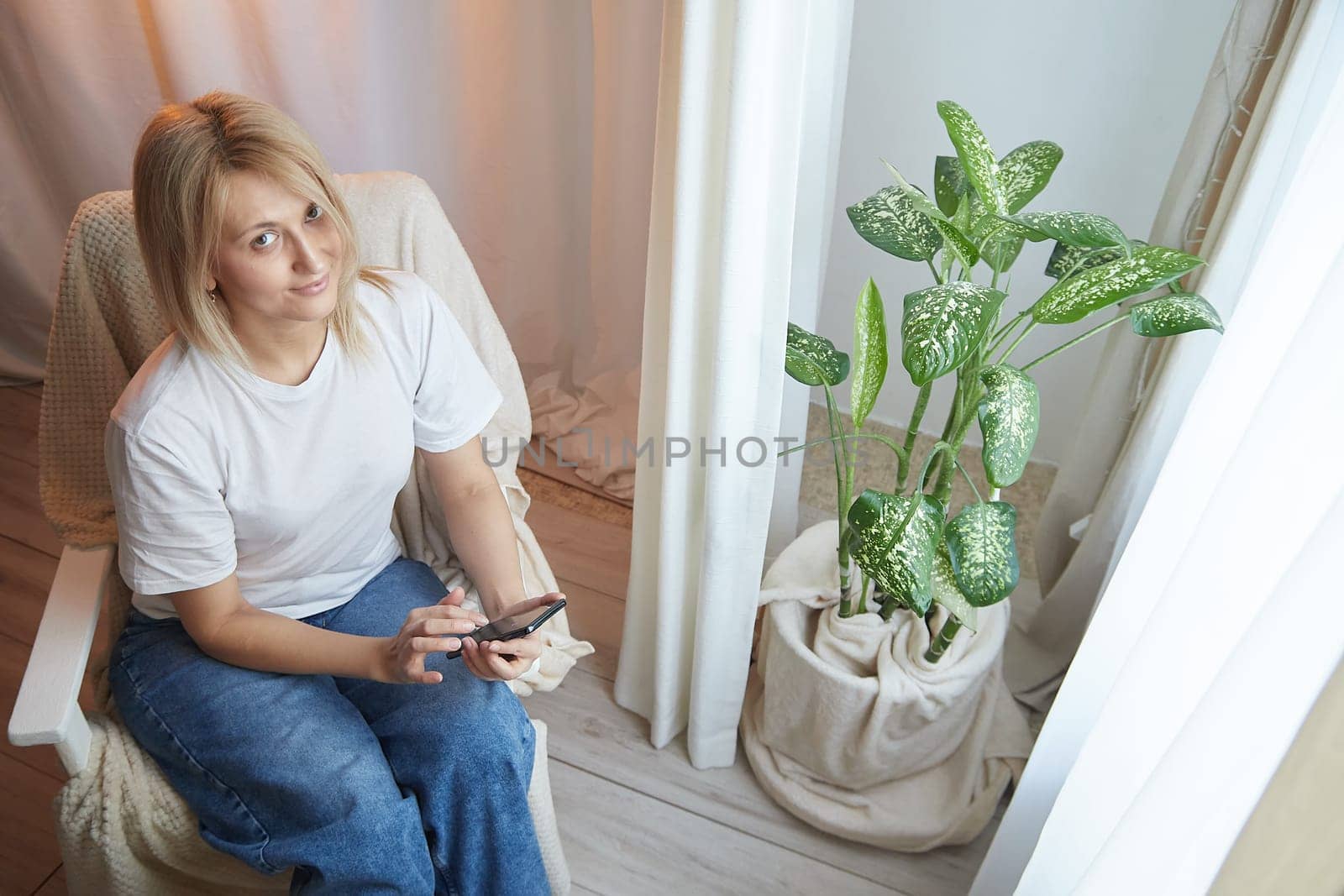 Young blonde woman using cell phone at home. Girl is resting, chating and looking in camera on smartphone near window at home. Nice atmosphere. The concept of a cozy home and communication by Internet by keleny