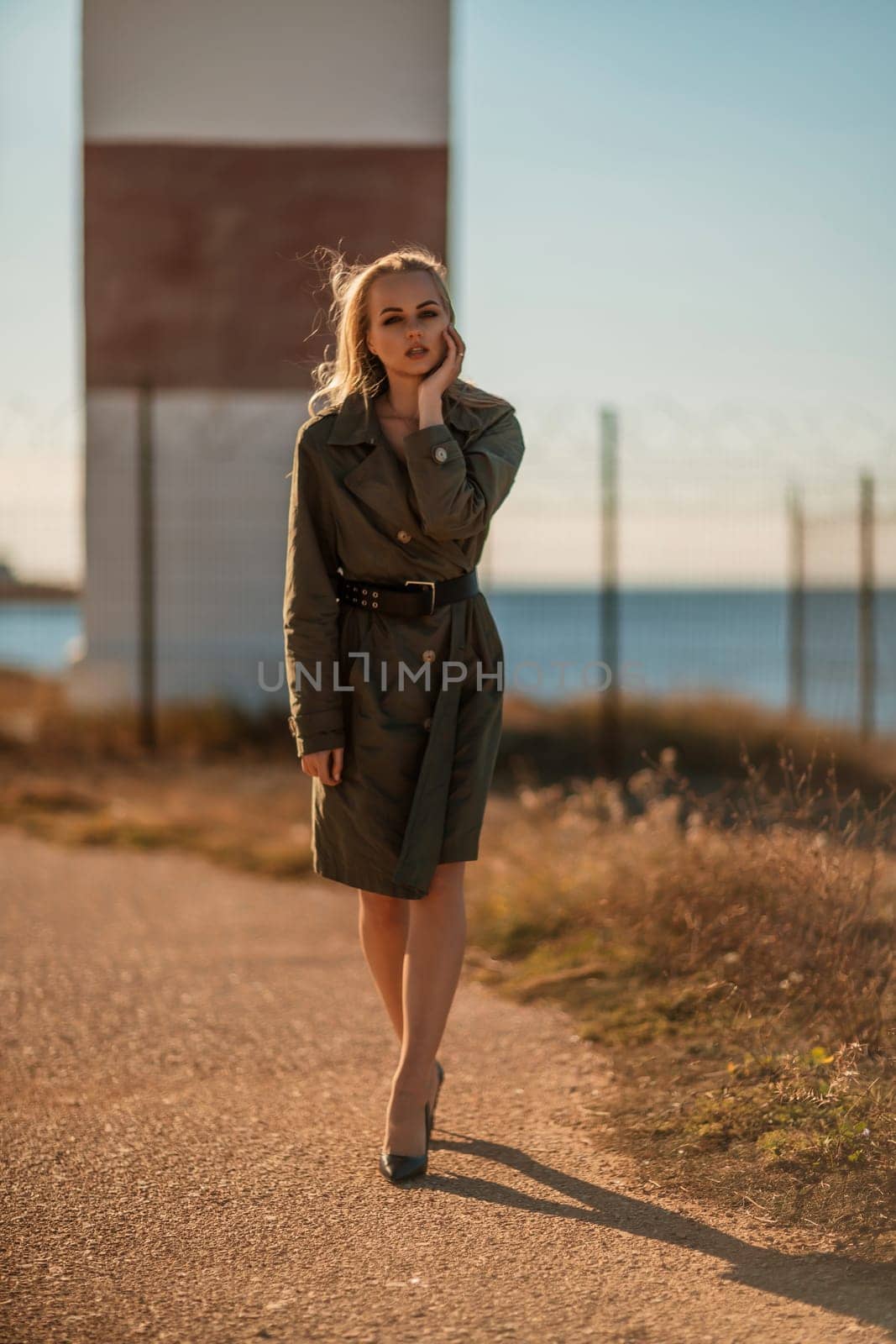 Portrait blonde sea cape. A calm young blonde in a khaki raincoat stands on the seashore against the backdrop of a lighthouse