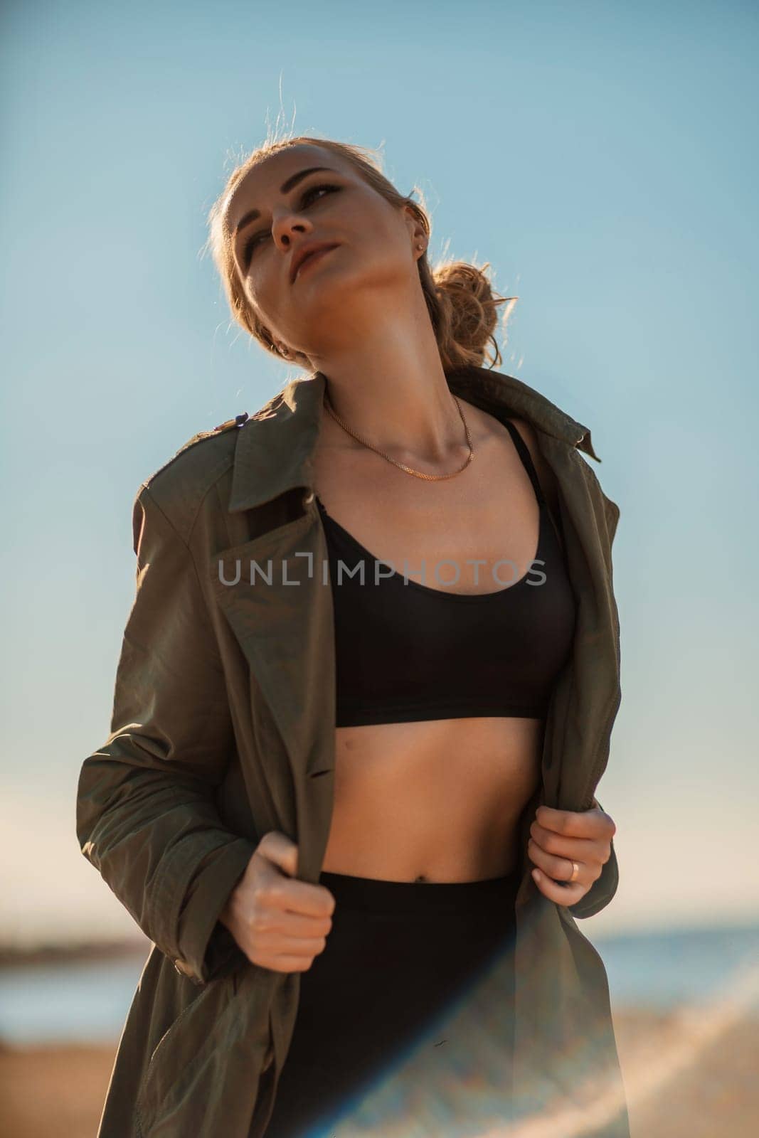 Portrait blonde sea cape. A calm young blonde in an unbuttoned khaki raincoat stands on the seashore, under the raincoat there is a black skirt and top.