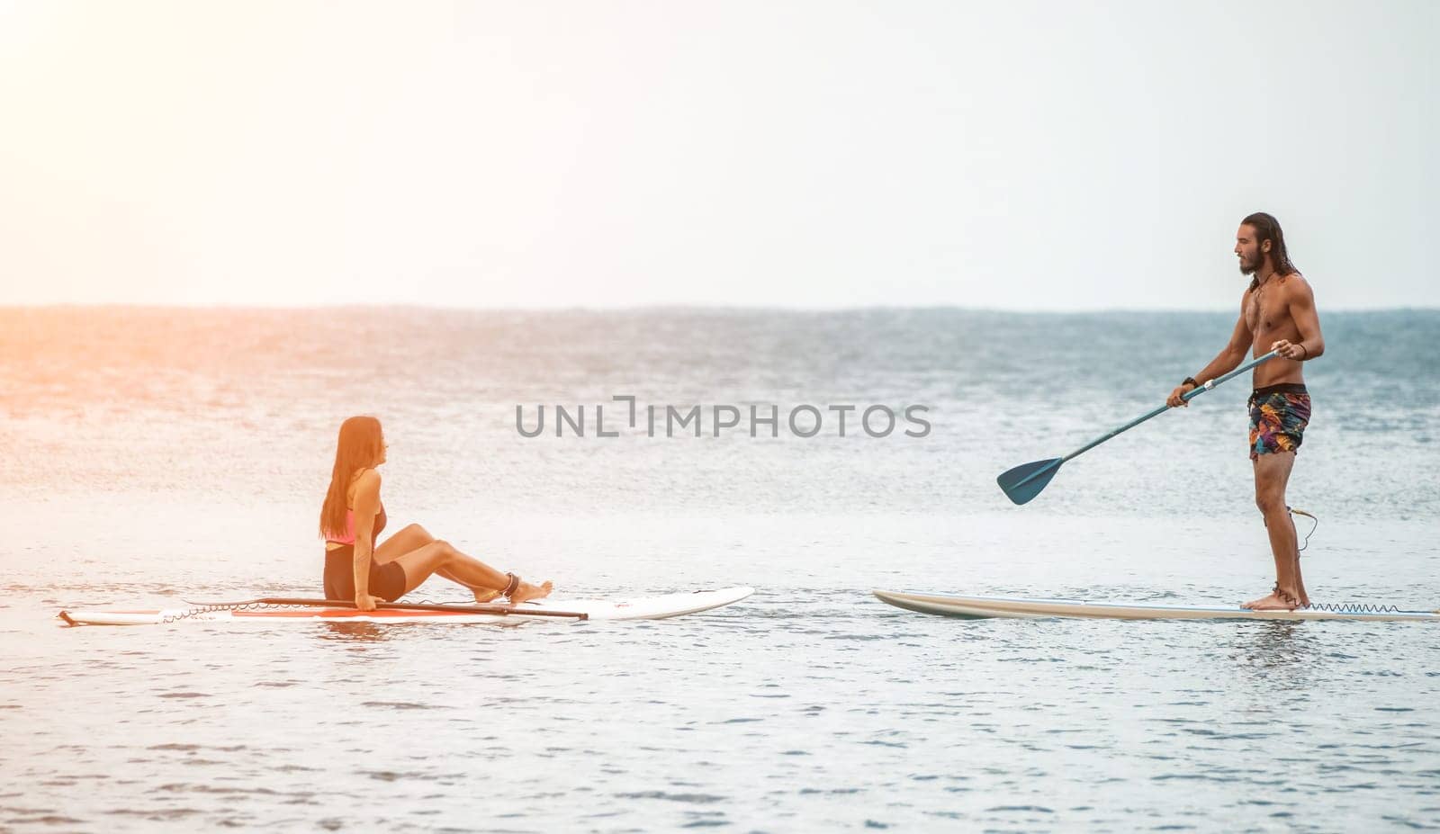 Sea woman and man on sup. Silhouette of happy young woman and man, surfing on SUP board, confident paddling through water surface. Idyllic sunset. Active lifestyle at sea or river