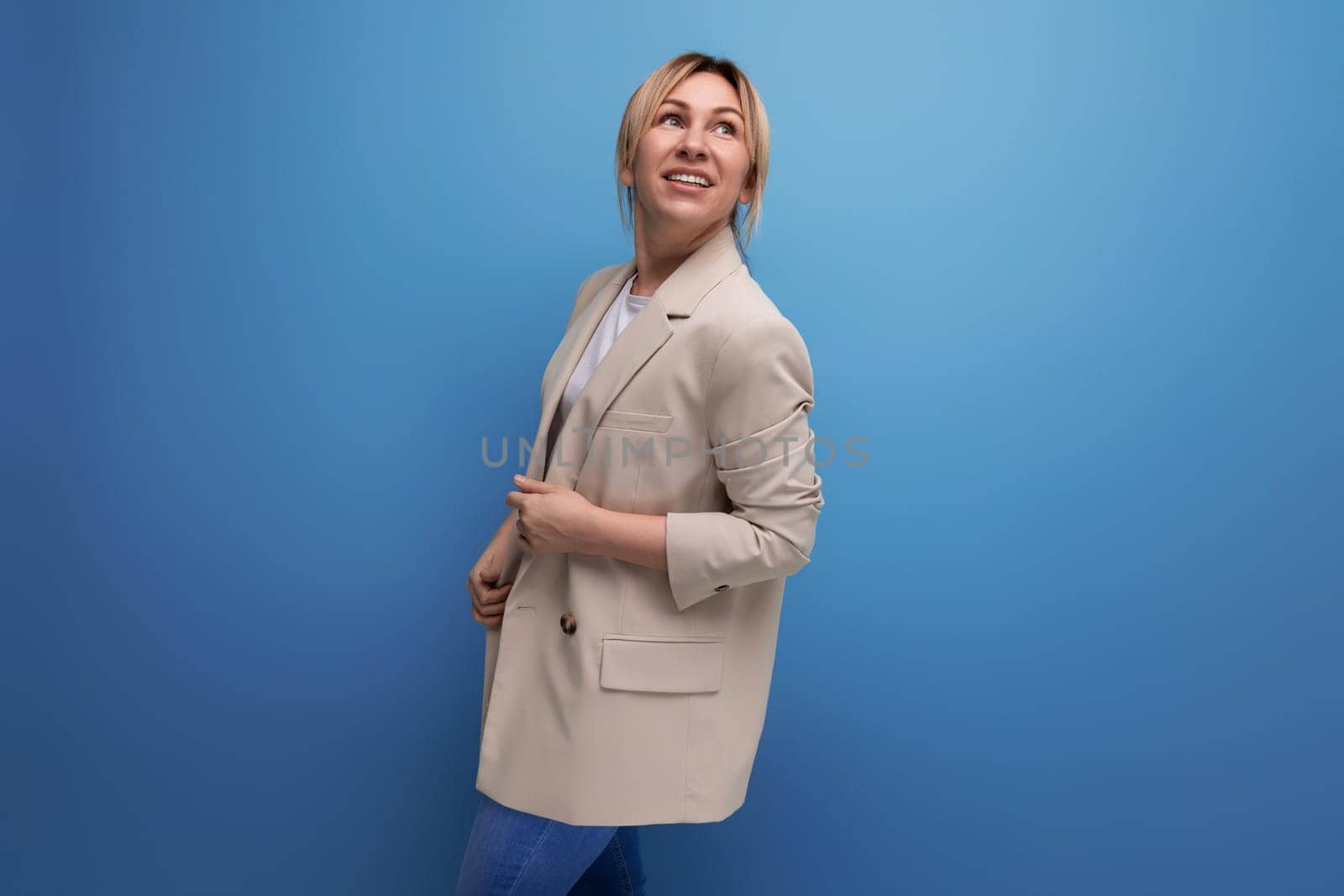 well-groomed blonde millennial woman in a beige jacket posing confidently on a blue background.