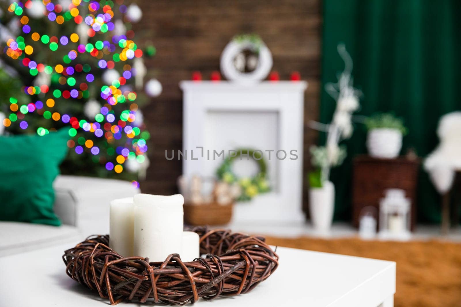 Close-up view of a few candles set in the center of a garland woven from brown wicker. A Christmas tree with lights and a fireplace stand in the blurry background.