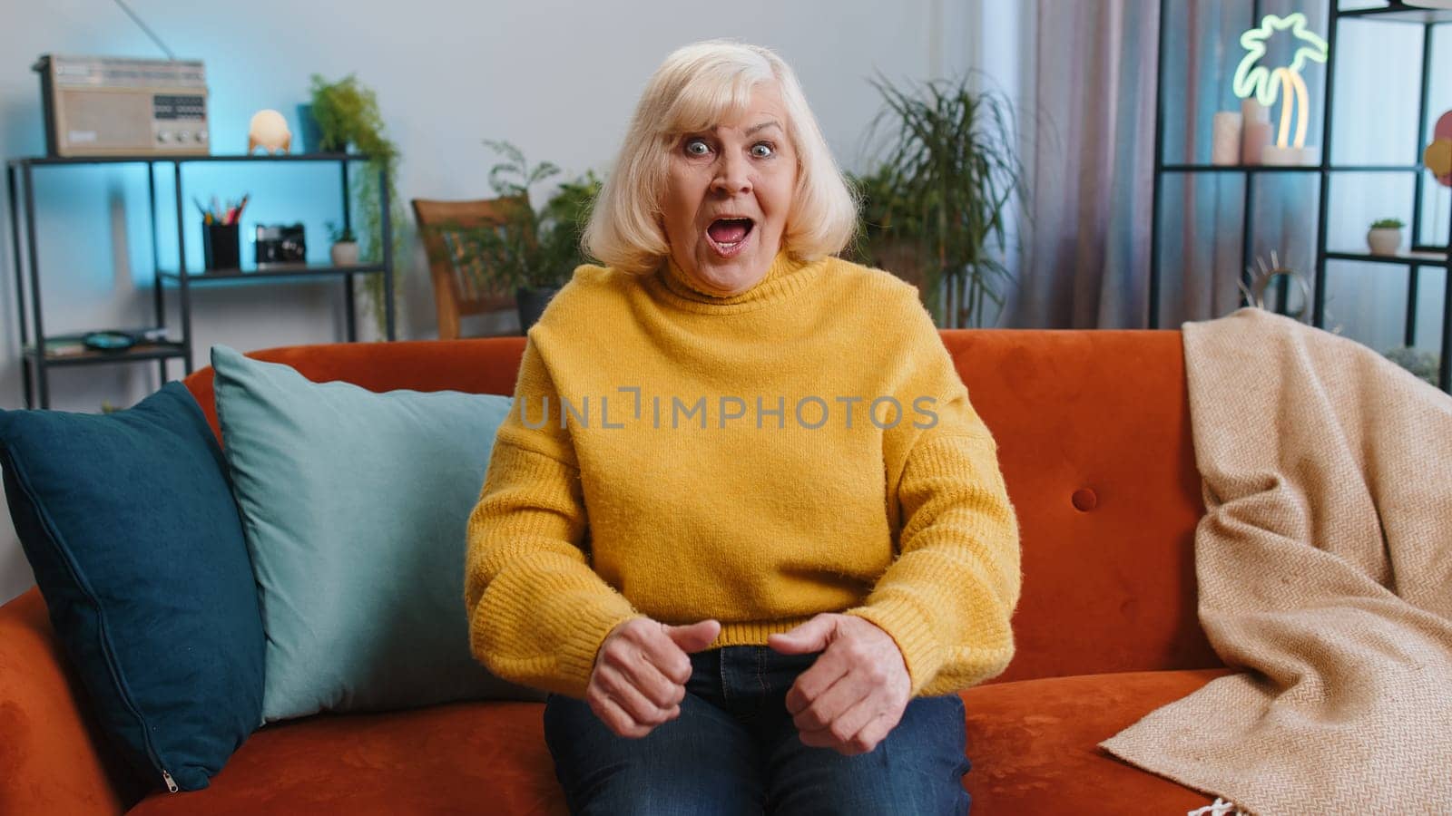 Excited senior grandmother woman on couch living room watching interesting TV serial sport game, film online social media movie content at home apartment. Elderly old pensioner enjoying entertainment