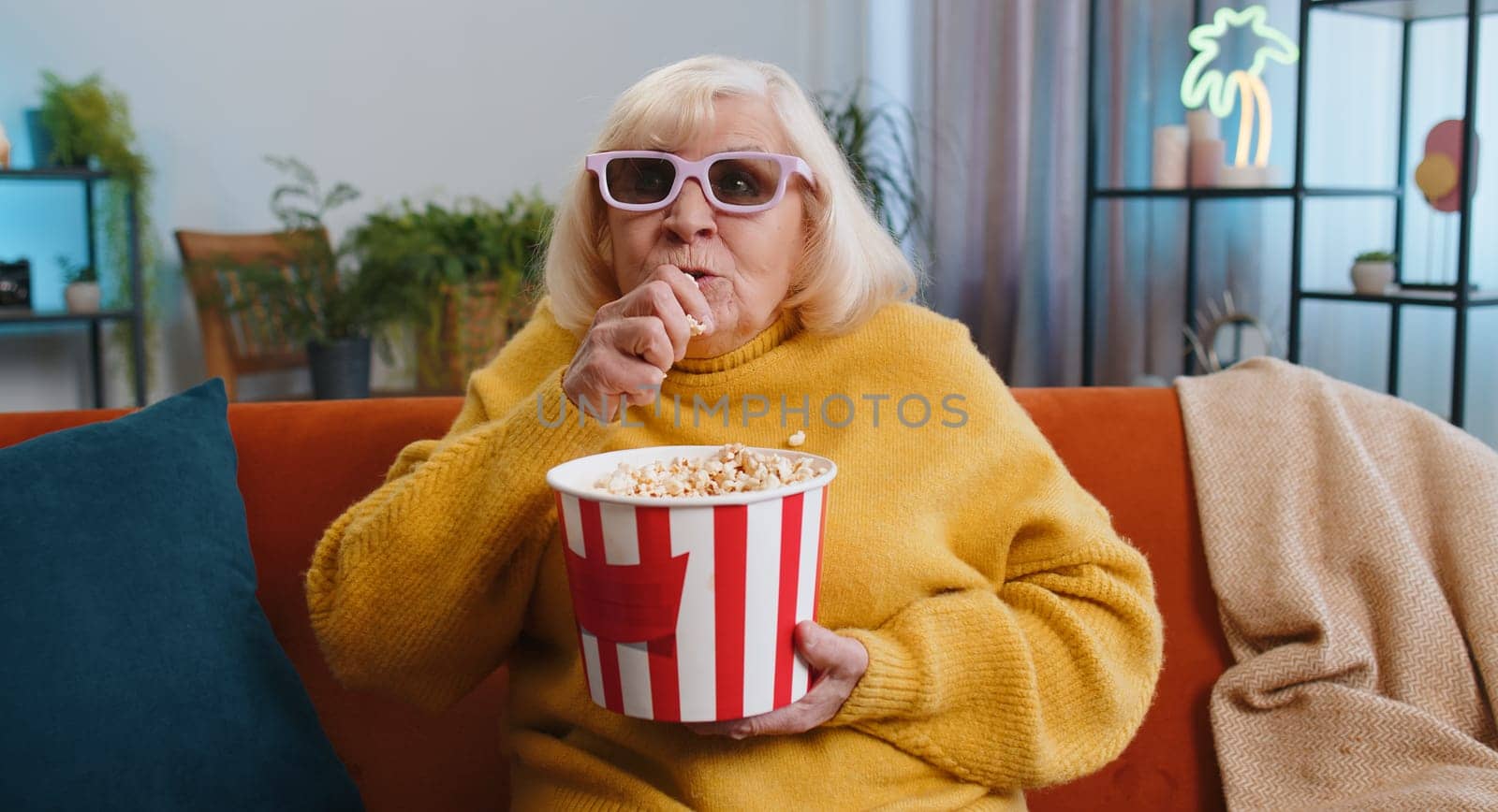 Senior old woman in 3D glasses on couch eating popcorn snacks and watching interesting TV serial sport game film online social media movie content at home apartment. Grandmother enjoying entertainment