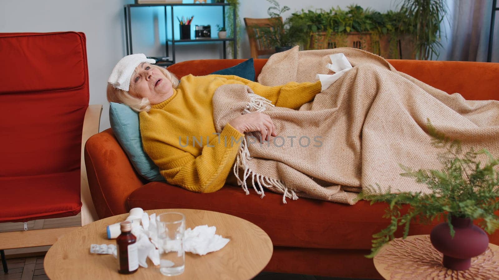 Grandmother ill woman suffering from cold or allergy lying on sofa in room at home apartment. Sick senior lady with towel on head blows sneezes wipes snot into napkin. Coronavirus quarantine, pandemic