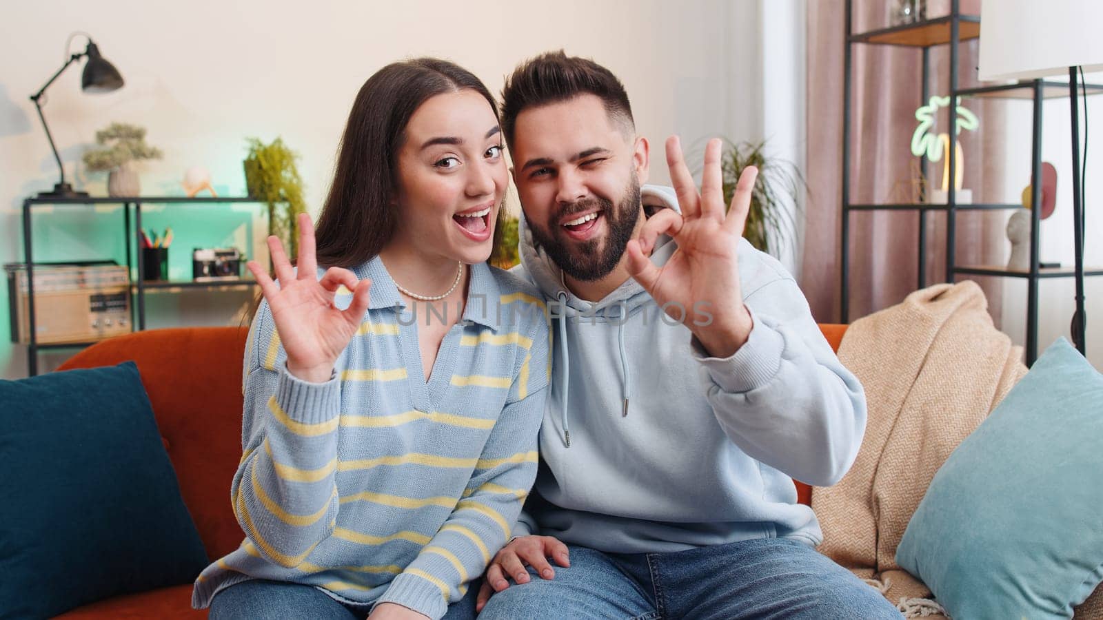 Okay. Happy cheerful family married couple man woman looking approvingly at camera showing ok gesture, positive like sign, approve something good at home. Young husband wife together on sofa in room