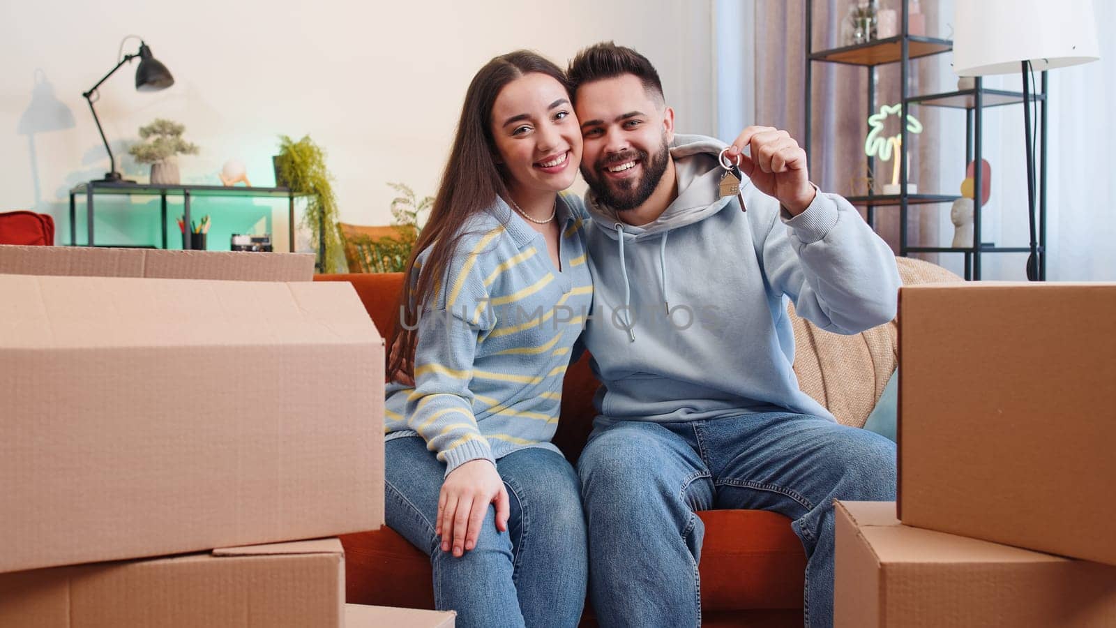 Young family marriage couple man woman lifting hand showing the keys of new home house apartment, buying or renting real estate property, mortgage loan at home. Husband wife together on sofa in room