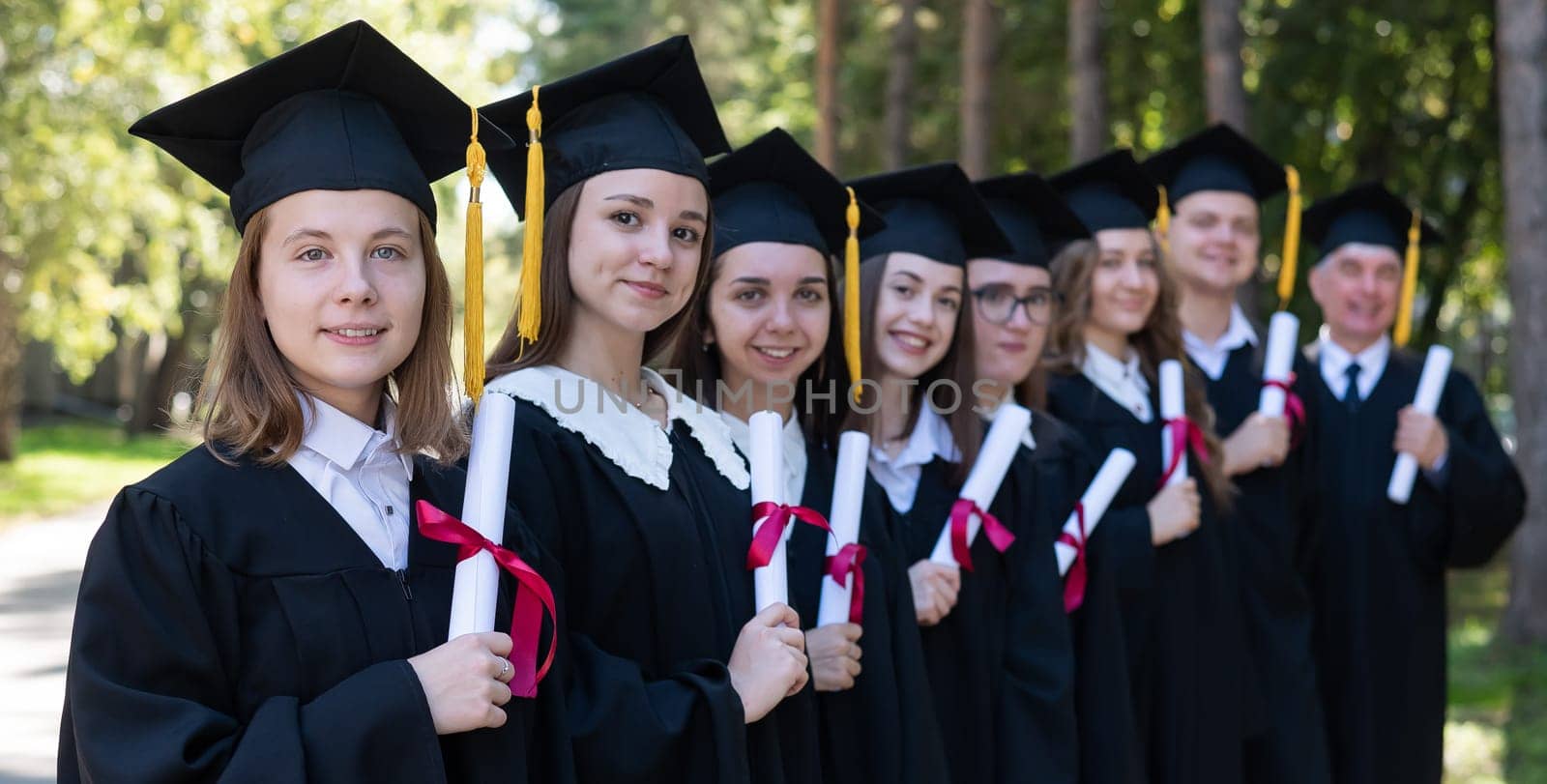 Row of young people in graduation gowns outdoors. Age student