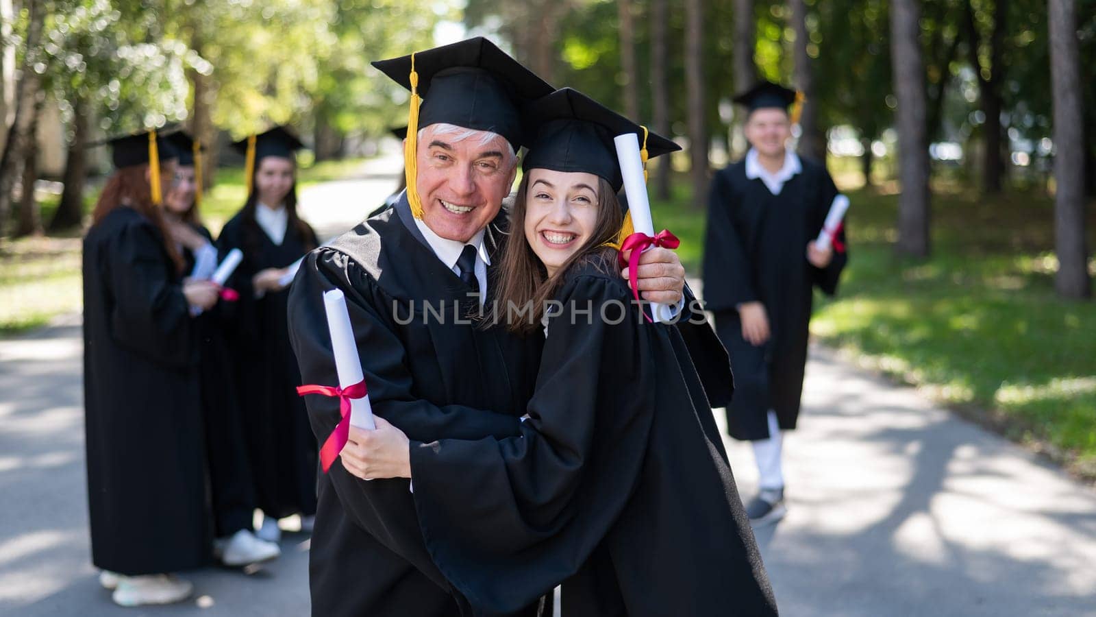 A group of graduates in robes outdoors. An elderly man and a young woman congratulate each other on their graduation