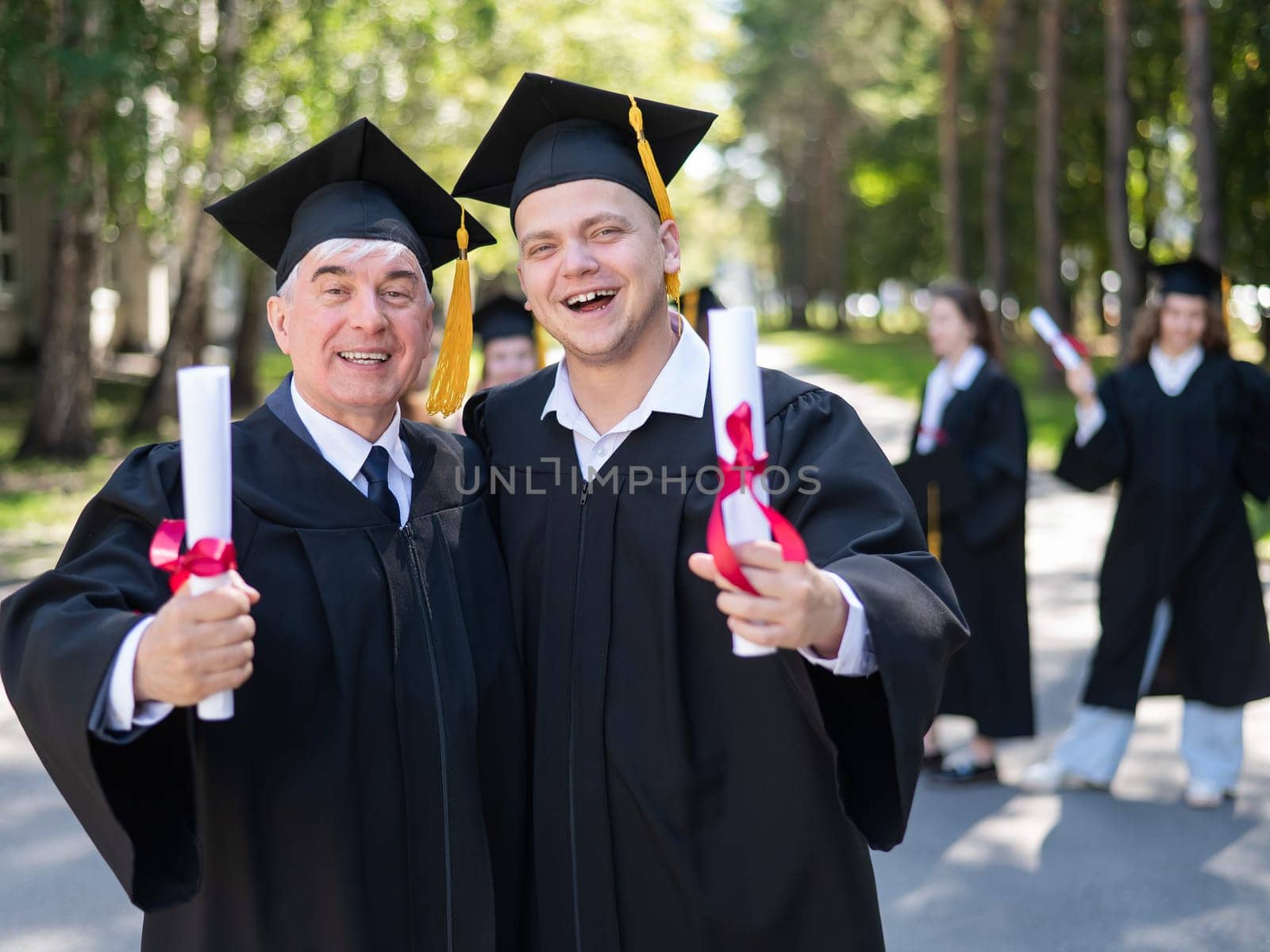 A group of graduates in robes outdoors. An elderly man and a young guy congratulate each other on receiving a diploma