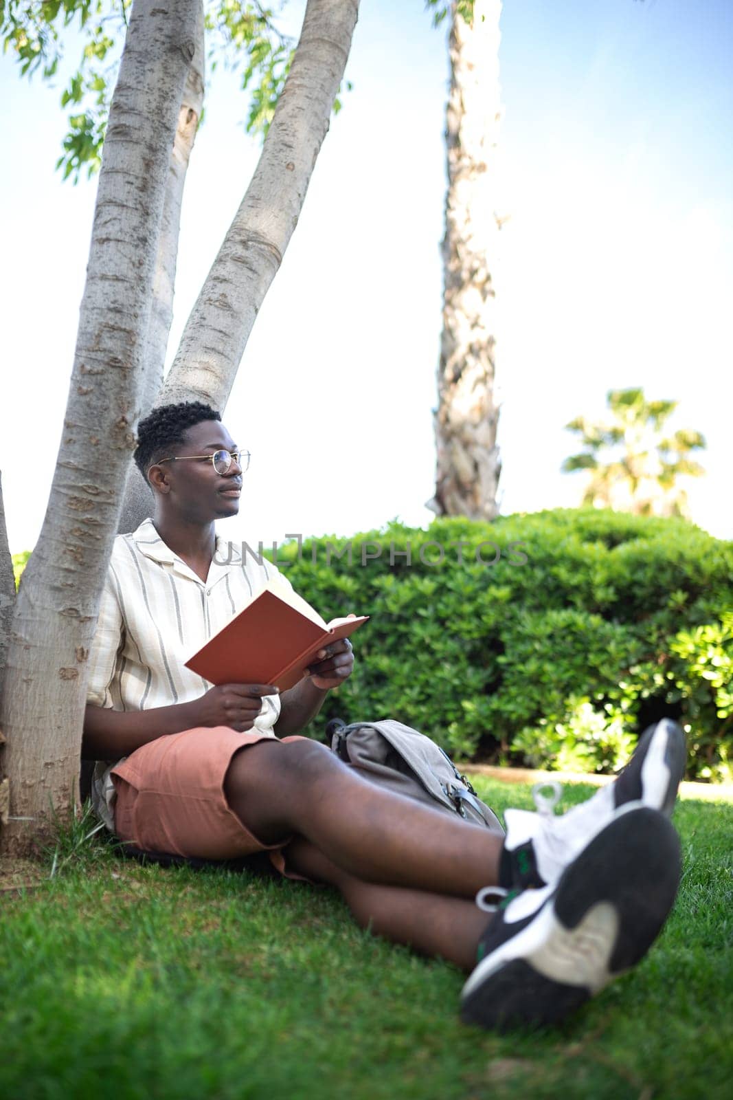 African american university student relaxing on campus.Young black man reading book outdoors. Copy space. by Hoverstock