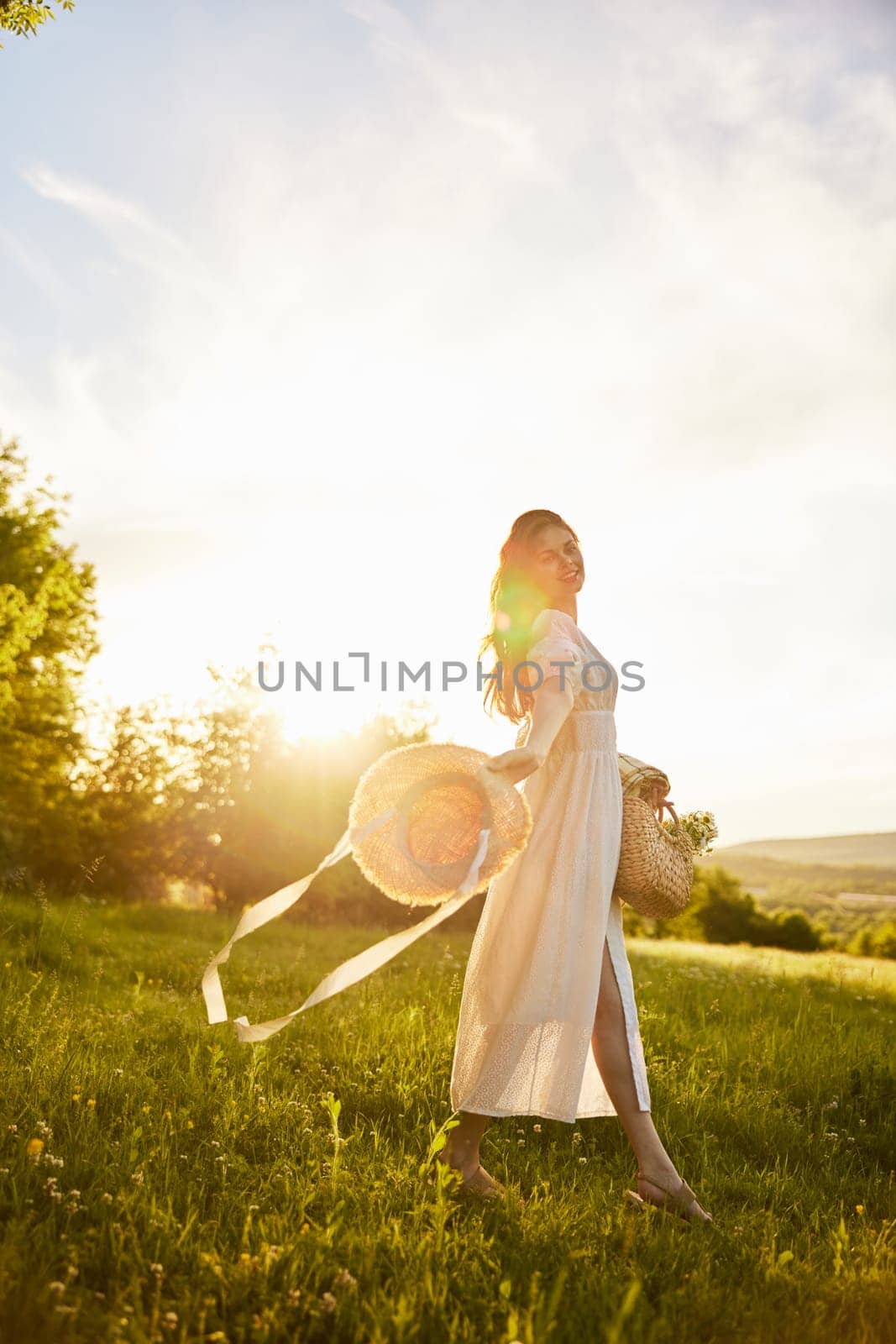 a woman in a light dress walks in nature holding a hat in her hands lit from the back by the sun. High quality photo