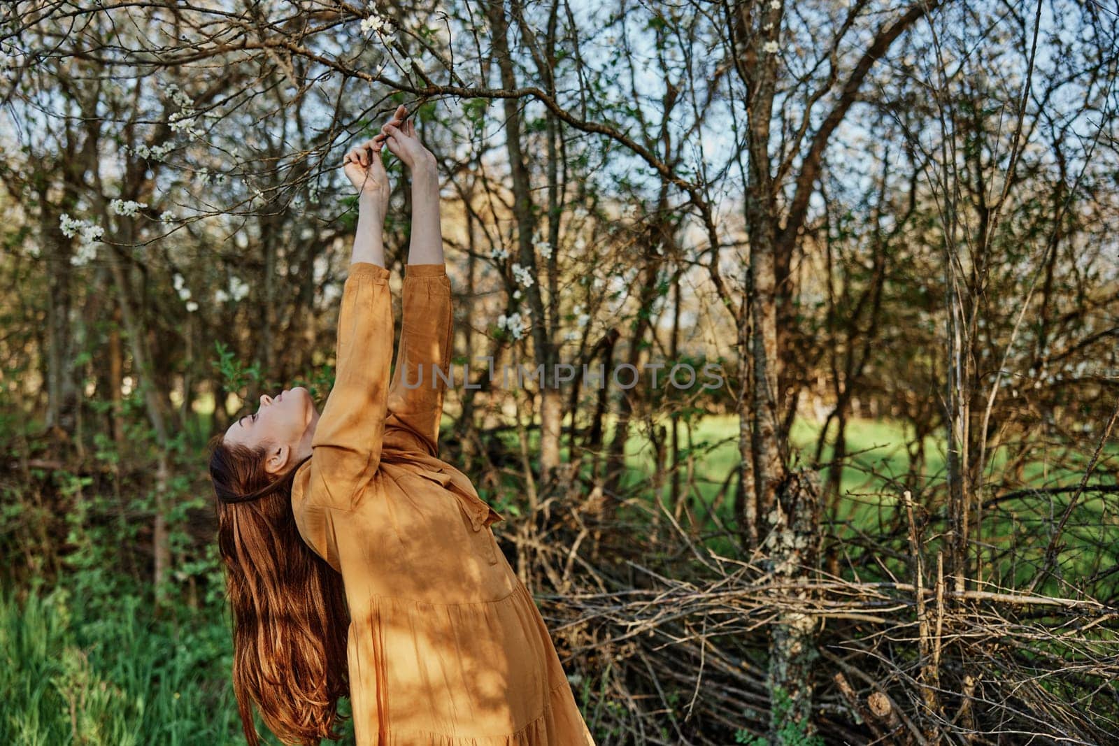 a beautiful, slender woman with long hair walks in the shade near the trees, dressed in a long orange dress, enjoying the weather and the weekend, joyfully raising her hands up. High quality photo