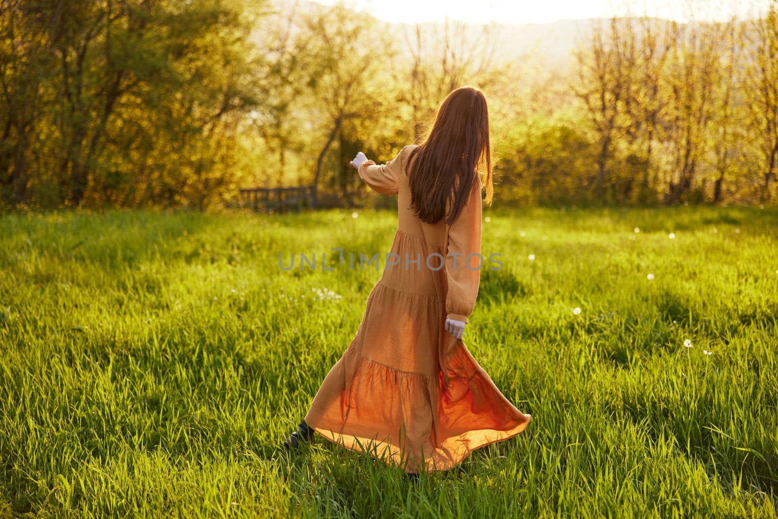 a relaxed slender woman enjoys the sunset standing in a green field with tall grass in an orange dress with her back to the camera, in warm summer weather. Horizontal photo. High quality photo
