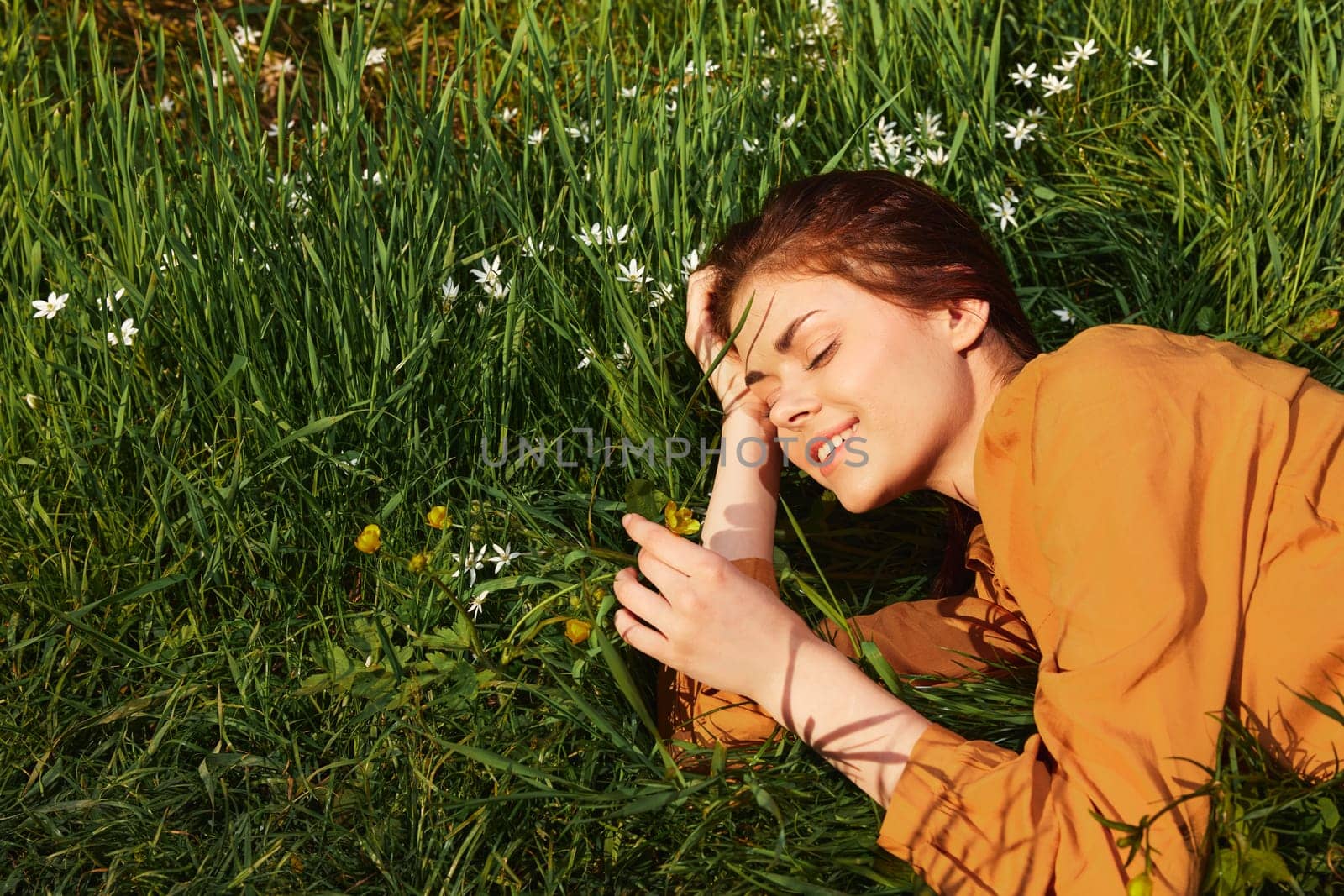 a calm woman with long red hair lies in a green field with yellow flowers, in an orange dress, smiling pleasantly, closing her eyes from the bright summer sun, resting her head on her hands by Vichizh