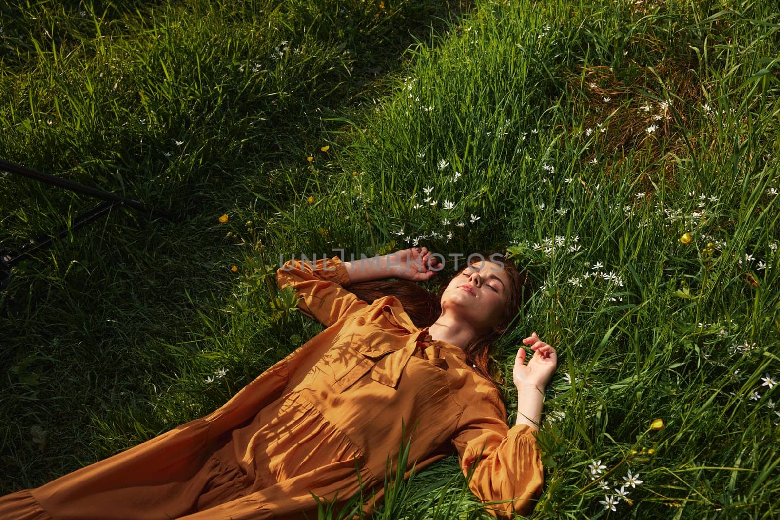 a happy, relaxed woman, resting lying in the green grass, in a long orange dress, with her eyes closed and a pleasant smile on her face, holding her hands near her face, enjoying harmony with nature and recuperating. High quality photo