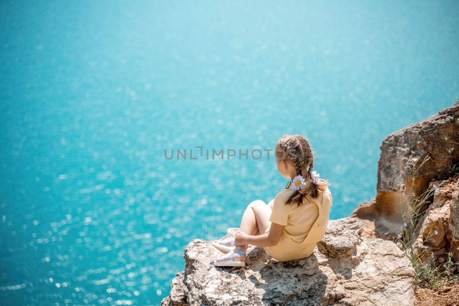 Happy girl perched atop a high rock above the sea, wearing a yellow jumpsuit and braided hair, signifying the concept of summer vacation at the beach