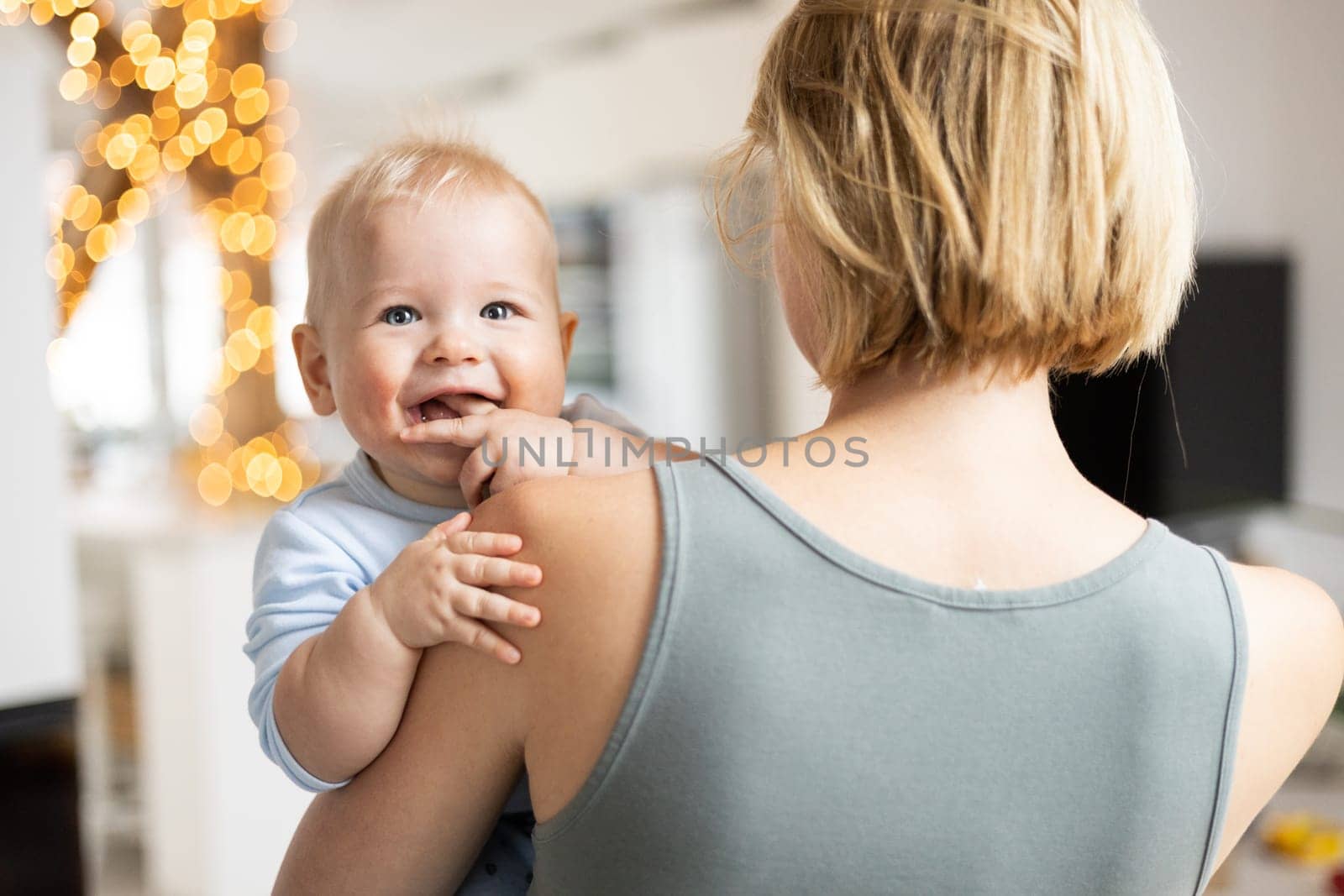 Blonde mother holding her adorable cheerful little infant baby boy. Cute infant child looking in camera and smiling at home. by kasto