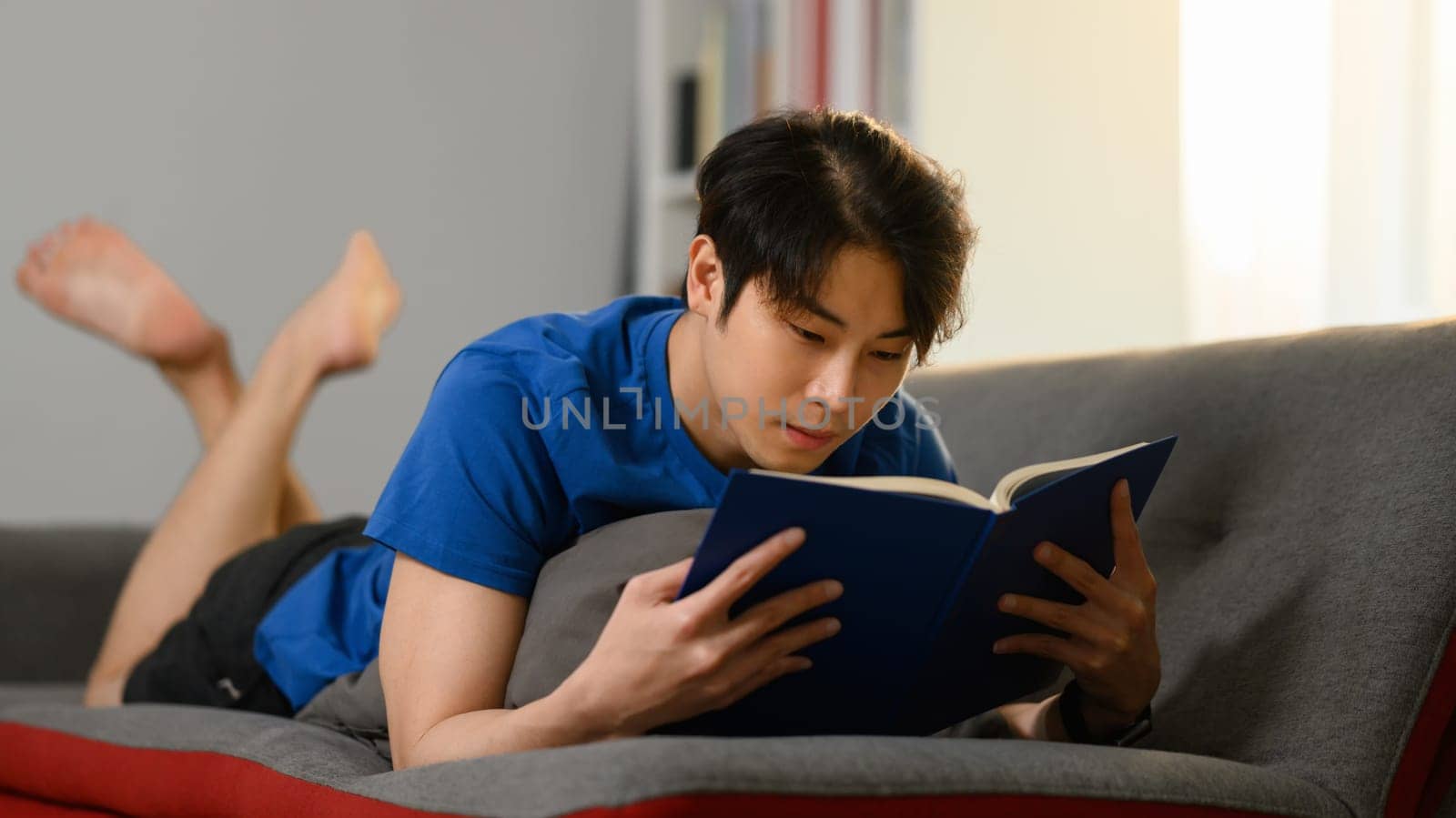 Relaxed asian man in casual clothes lying on couch and reading favorite novel. People, leisure and lifestyle concept.