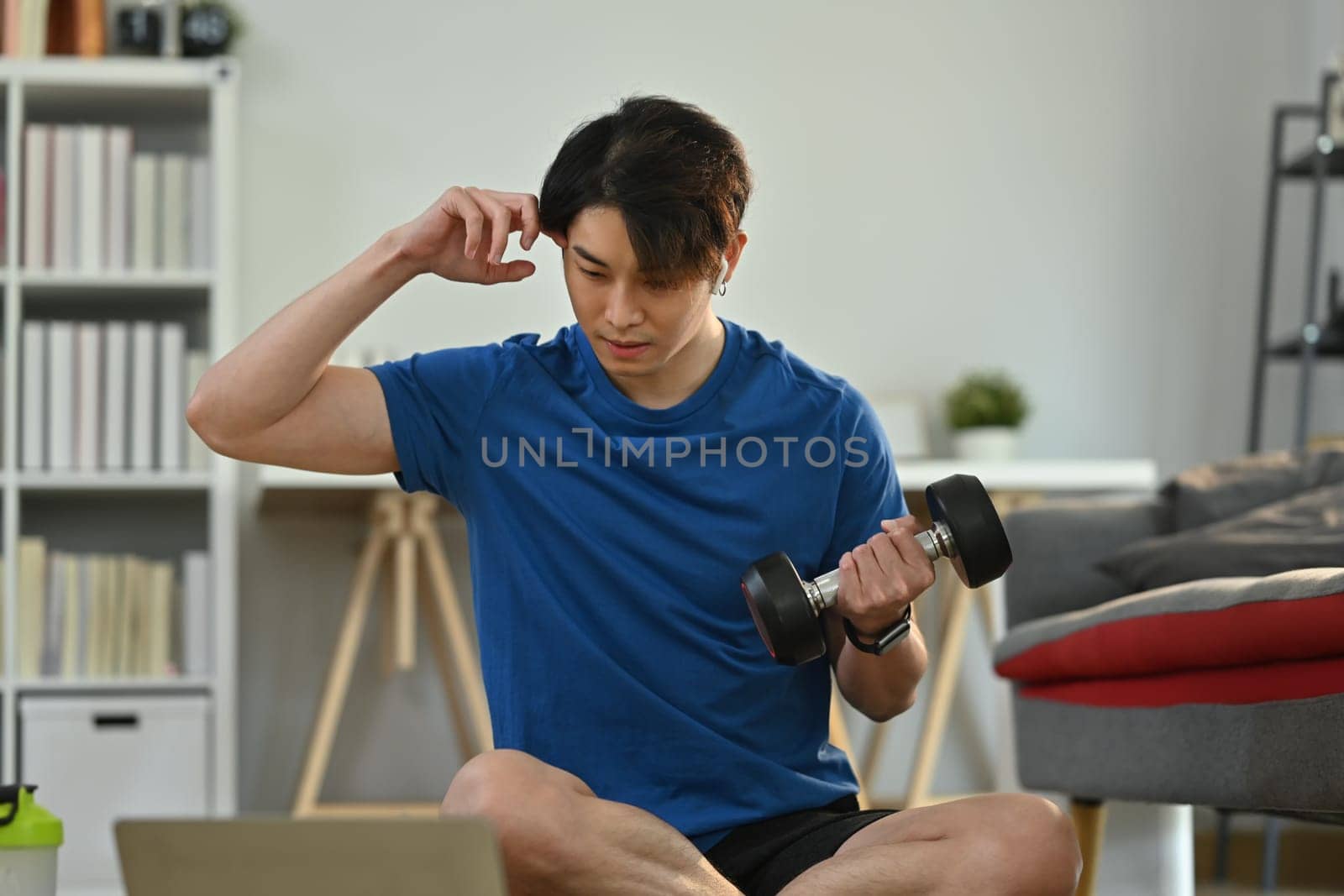 Active sportsman lifting dumbbells and watching video online training on laptop during morning workout at home by prathanchorruangsak