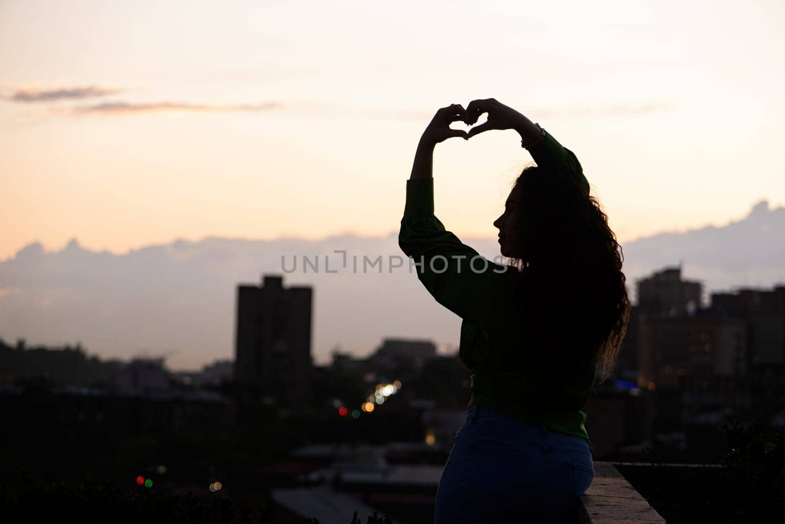 silhouette of beautiful woman on rooftop at urban sunset, making heart shape with hands