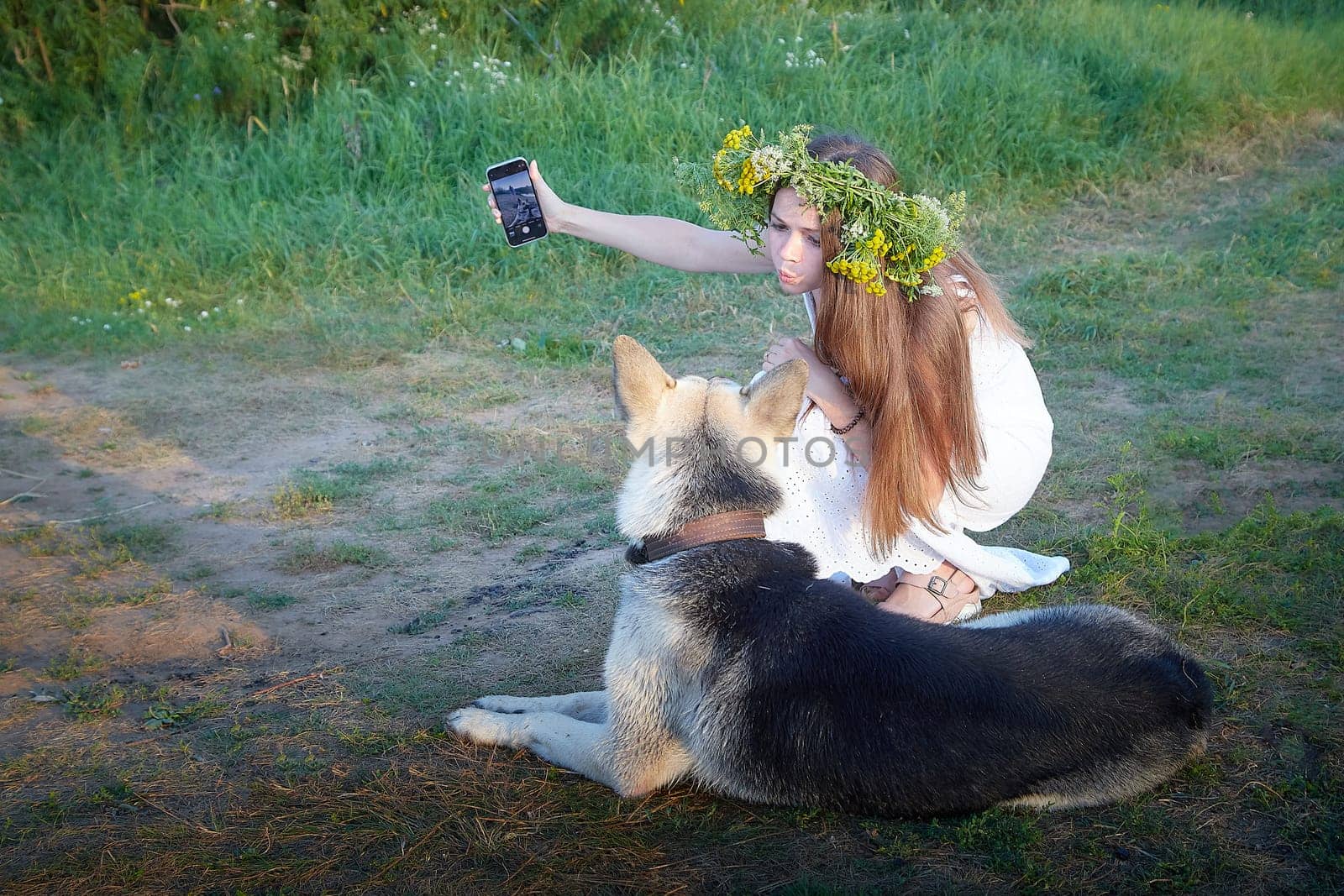 Young brunette girl in a white dress, sundress and wreath of flowers with big shepherd dog in summer on the coast of river or lake in the evening at sunset celebrating pagan holiday of Ivan Kupala