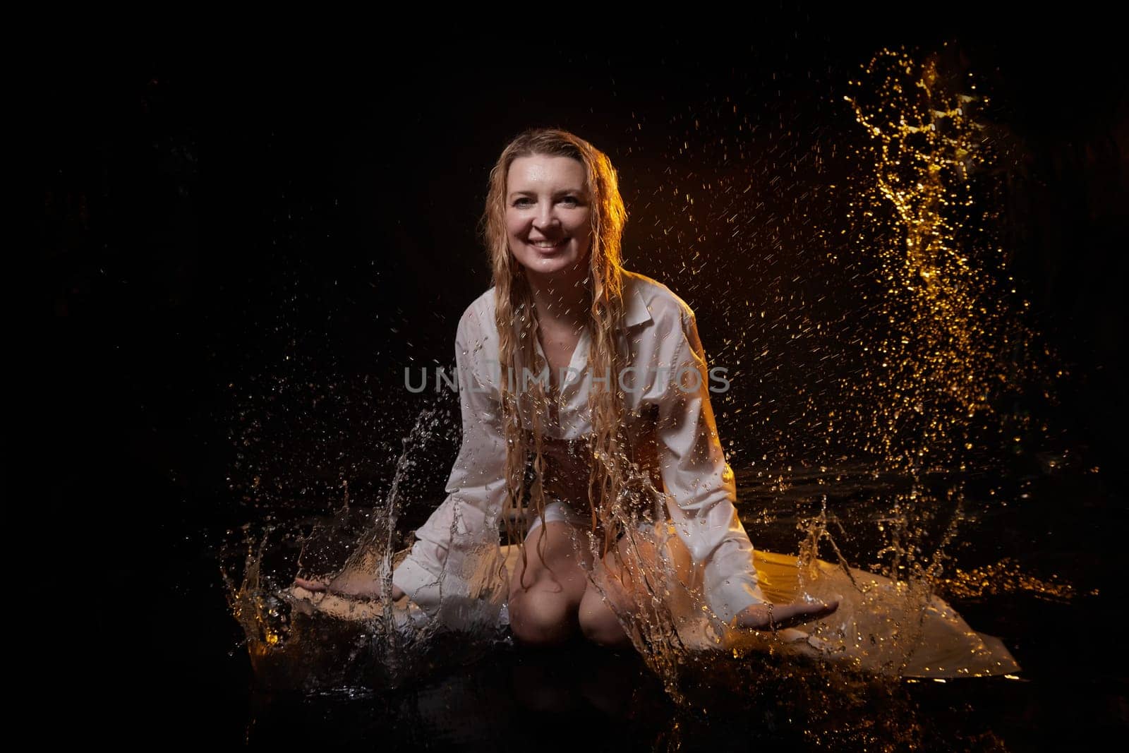 Beautiful sexy skinny girl in a dark hall with splashes of water. Female Model blonde woman posing with water on a black background in the studio by keleny