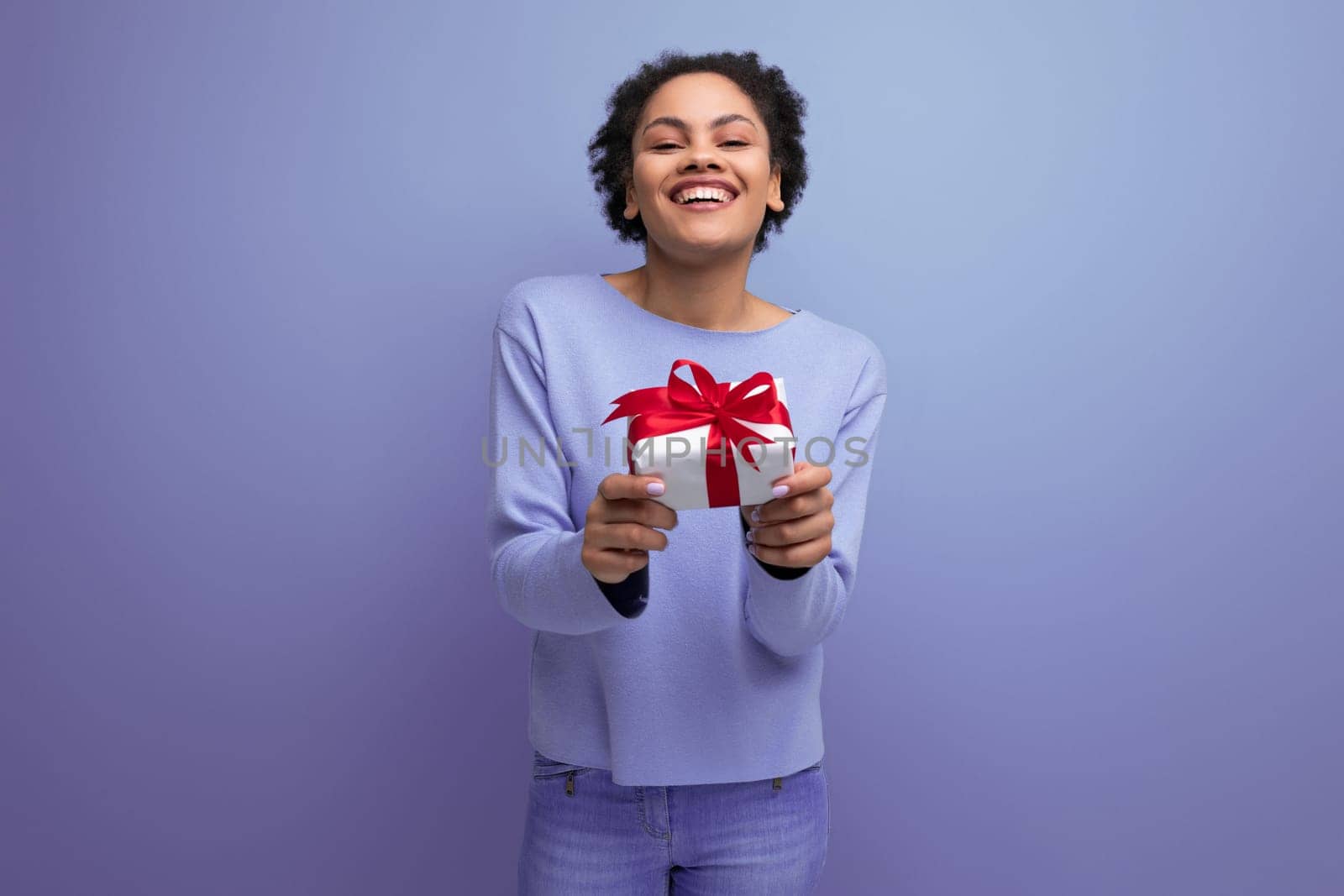 positive young brunette lady with afro curls holds out a gift box.