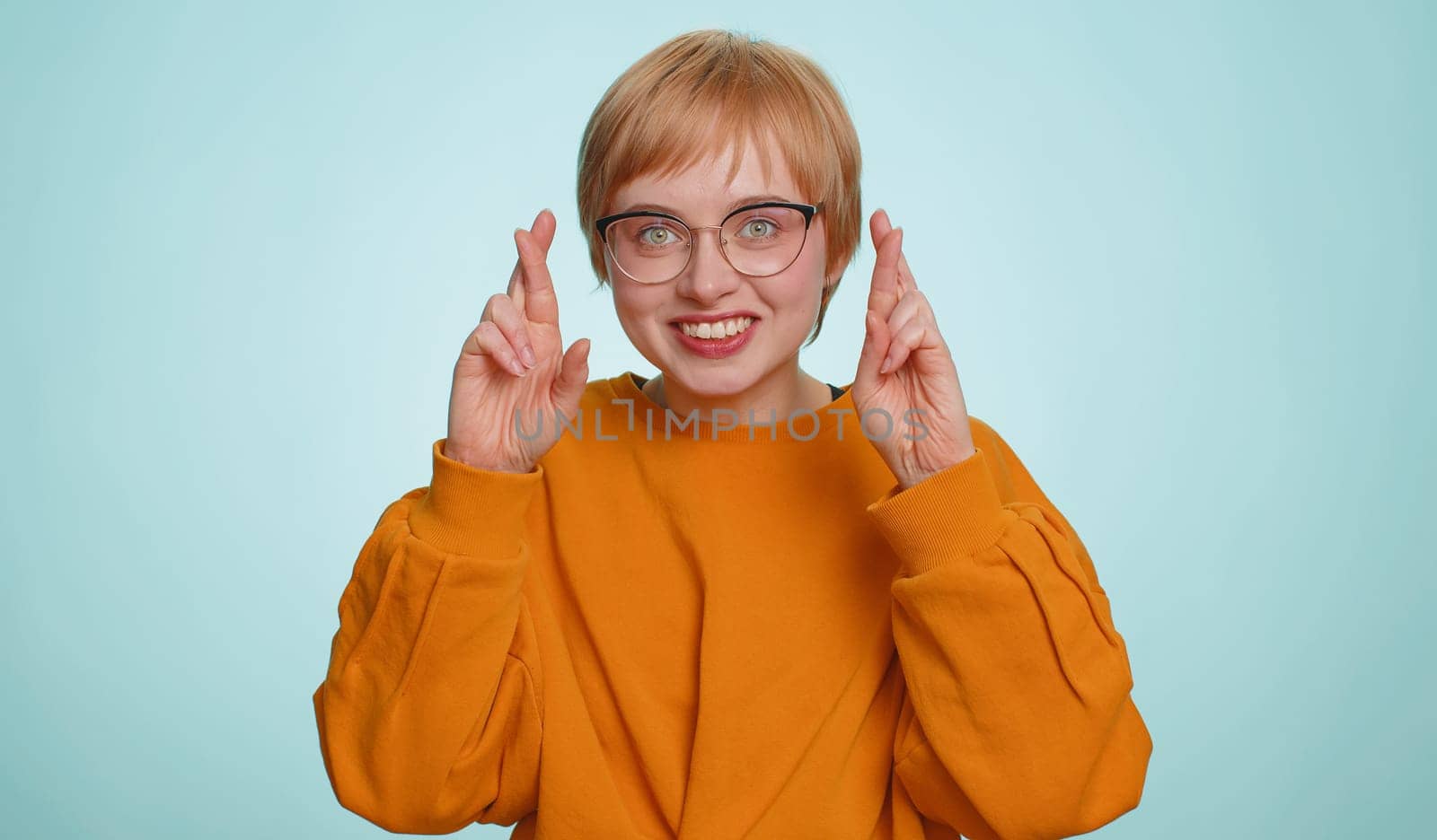 Excited blonde woman crossed fingers looking at camera asking for good luck news, wishing good exam results, dreaming about win victory in lottery jackpot. Girl isolated on yellow studio background