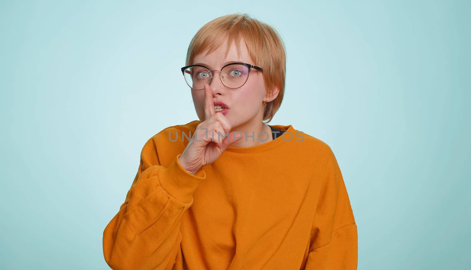 Shh be quiet please. Young woman presses index finger to lips makes silence gesture sign do not tells secret, stop gossip. Pretty short haired blonde girl isolated alone on blue studio background