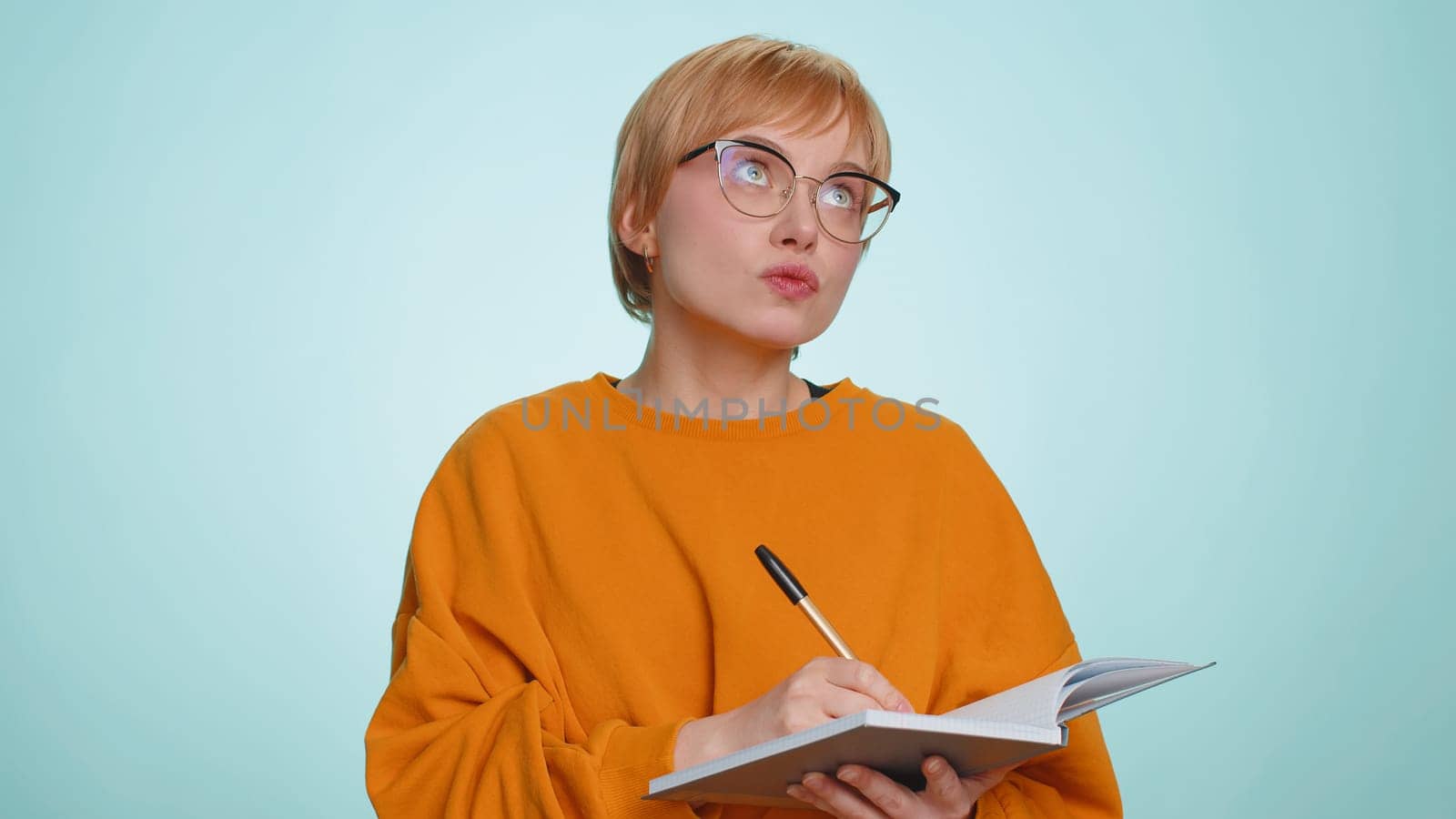 Thoughtful journalist woman making notes, writing down thoughts with pen into notepad notebook diary, to do list, good idea. Pretty short haired blonde girl isolated alone on blue studio background
