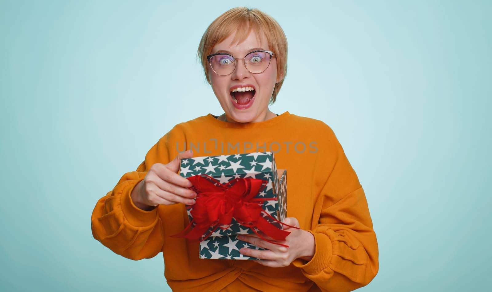 Woman in glasses opening gift box, unwrapping birthday surprise and expressing great happiness amazement, satisfied with best present. Blonde short haired girl isolated on blue studio background