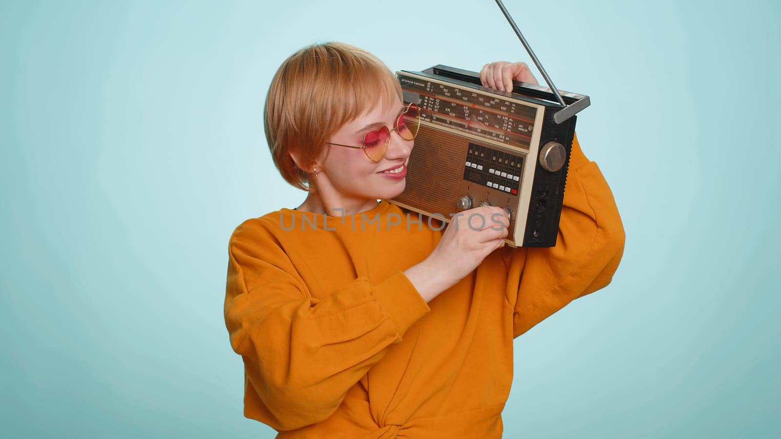 Woman in sunglasses using retro tape record player to listen music, disco dancing of favorite track, having fun, entertaining, fan of vintage technologies. Blonde girl isolated on blue background