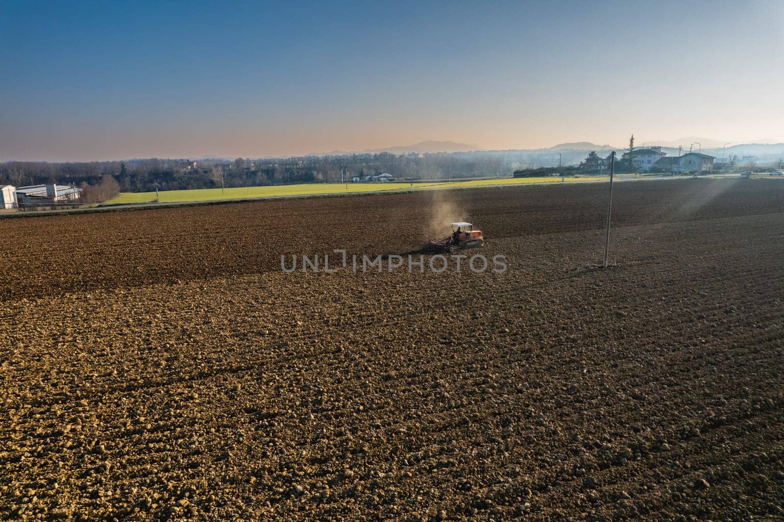 farmer driving a crawler tractor drags a cloud of dust behind, ploughing, power harrowing surface crust breaking to facilitate the birth and seeding of plants on dry soil