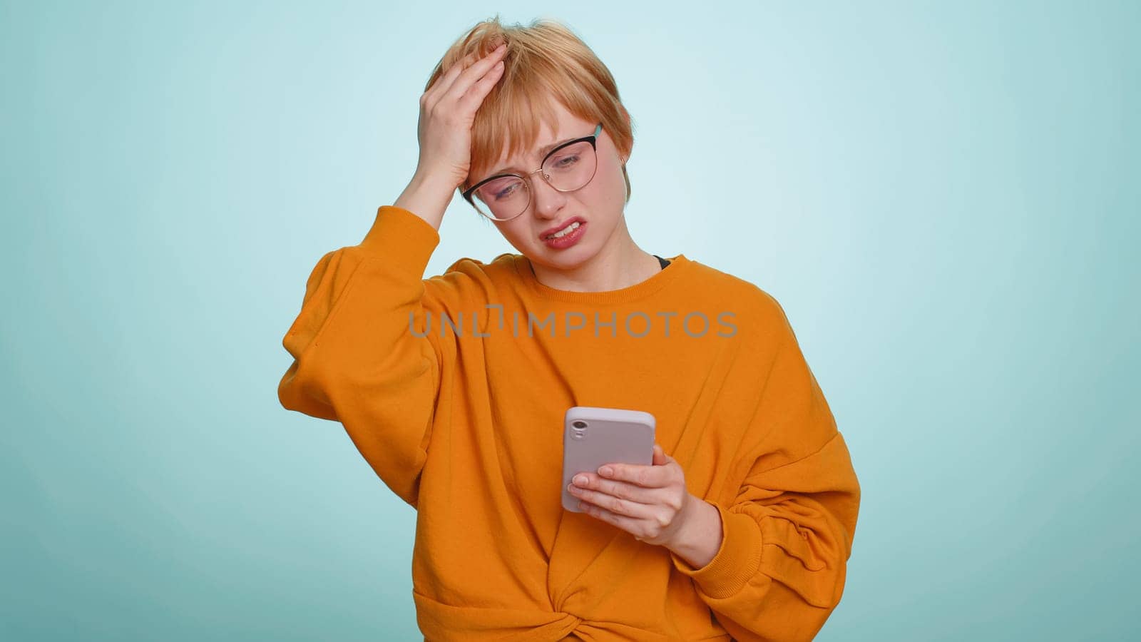 Woman use mobile phone smartphone typing browsing, loses becoming surprised sudden lottery results, bad fortune, loss, fail. Pretty blonde girl isolated alone on blue studio wall background, indoors