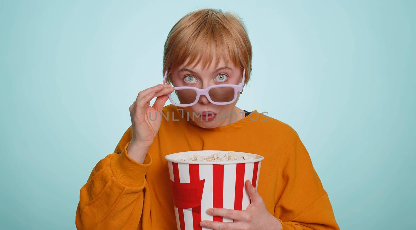 Excited young woman in 3D glasses eating popcorn and watching interesting tv serial, sport game, film, online social media movie content. Blonde short haired girl isolated on blue studio background