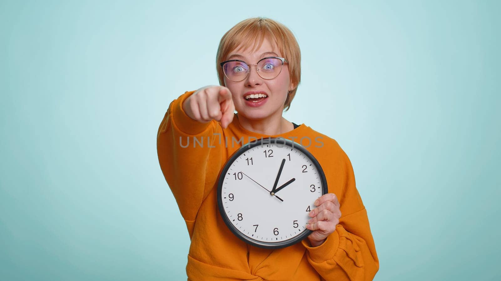 It is your time. Young woman in glasses showing time on wall office clock, ok, thumb up, approve, start, pointing finger at camera. Blonde short haired girl isolated on blue studio background, indoors