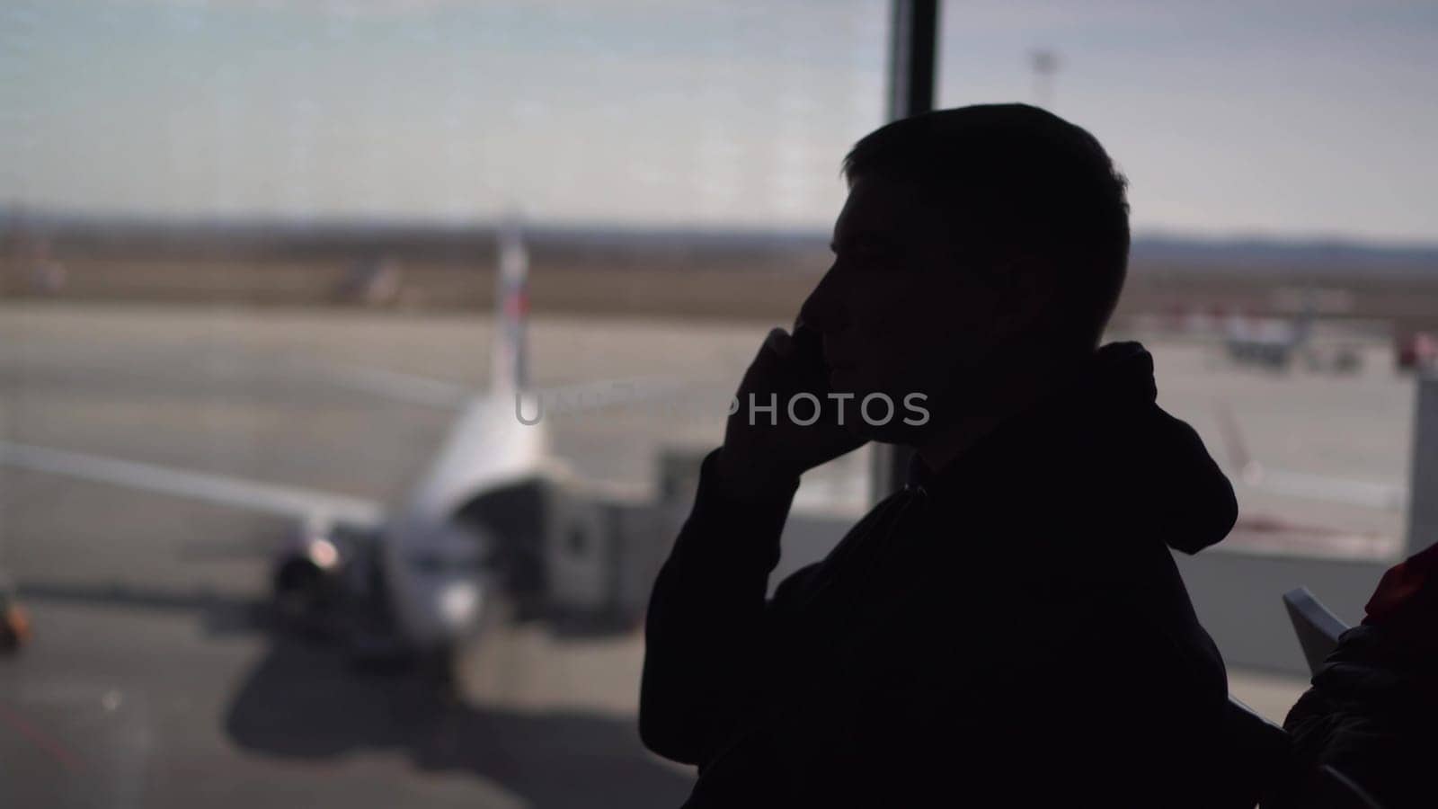 A young man in a sweatshirt is sitting at the airport and talking on the phone. Silhouette of a man on the background of the planes outside the window. 4k