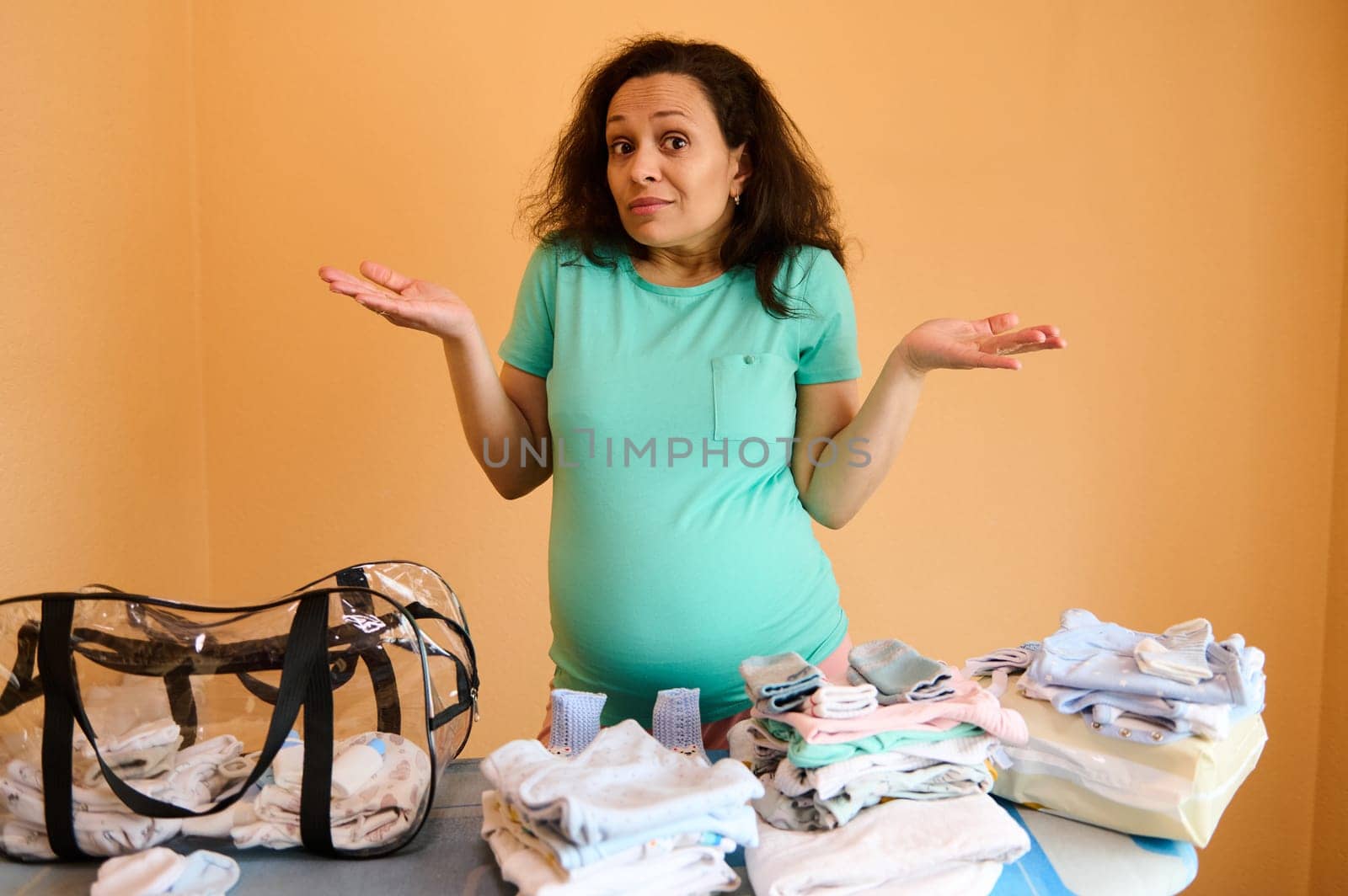 Surprised multi ethnic adult pregnant woman, standing by ironing board with clean ironed baby laundry, holding hands palms up, looking at camera, asking what to pack into a bag for maternity hospital