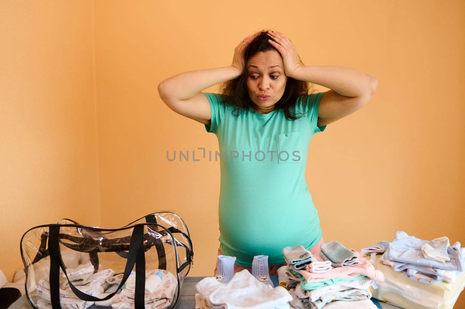Multitasking adult pregnant woman standing by ironing board with clean ironed newborn baby clothes, putting hands on head, feeling doubt to choose what things to pack into a bag for maternity hospital