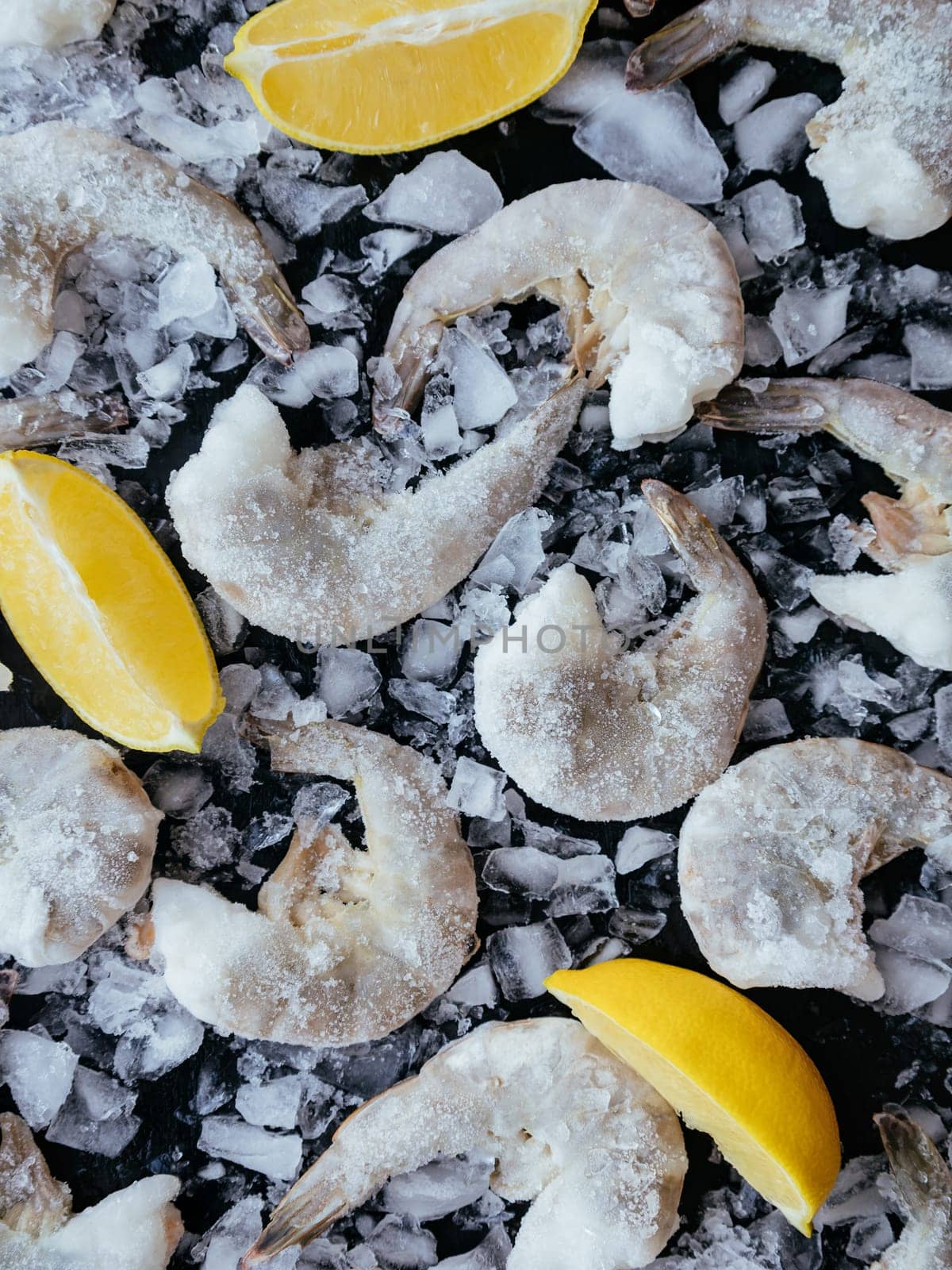 Seafood background for presentation your products. Frozen ocean jumbo shrimps with ice and lemon on black background for cooking. Pacific white shrimp or king prawns without head for meal preparation.