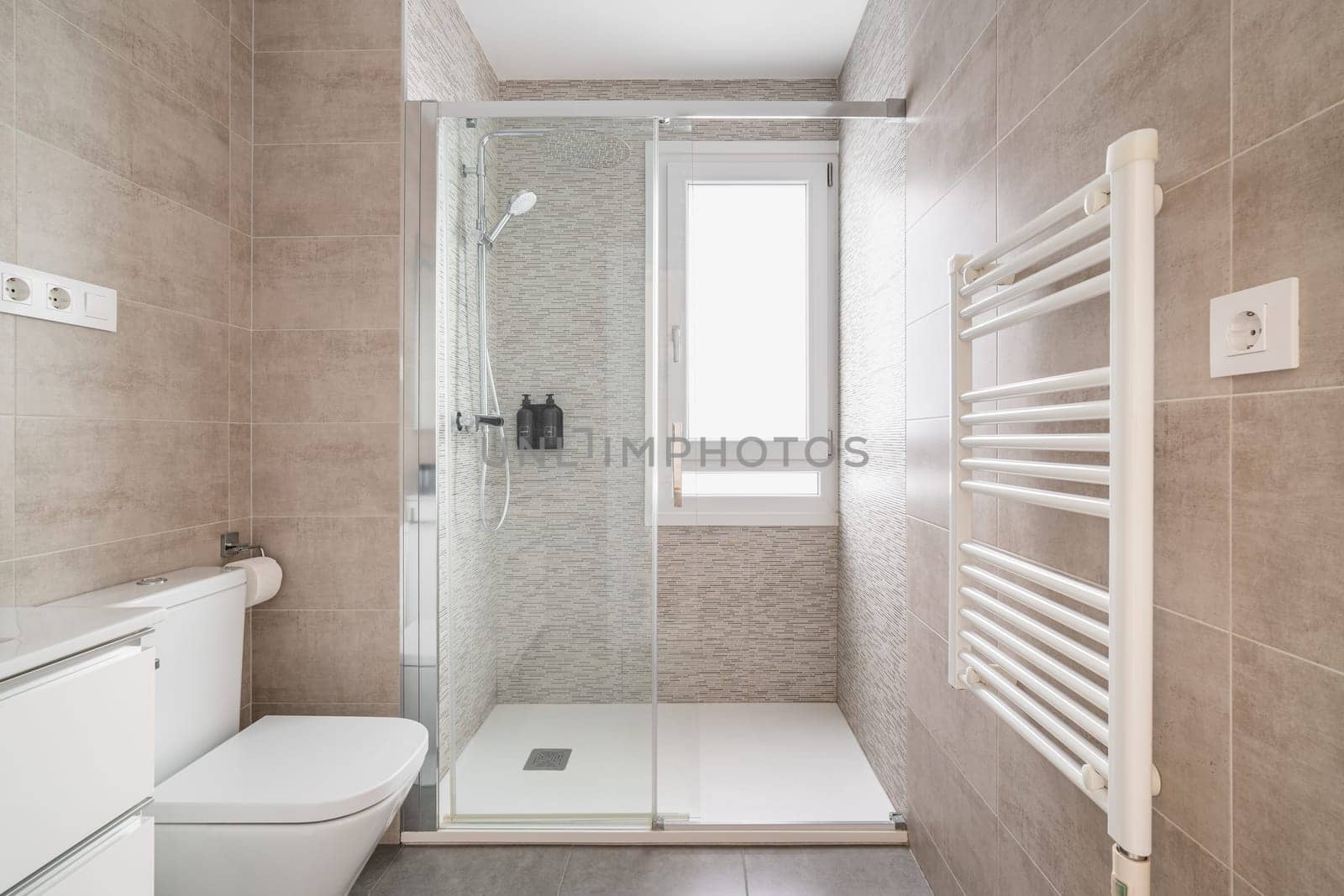 Comfortable bathroom with a toilet bowl and a shower cabin with tiles in beige tones and a window for natural light. The concept of a bathroom in a hotel or apartment after renovation.