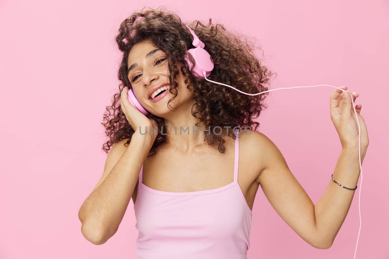 Happy woman wearing headphones with curly hair listening to music and singing along with her eyes closed in a pink T-shirt and jeans on a pink background DJ party, copy space. High quality photo