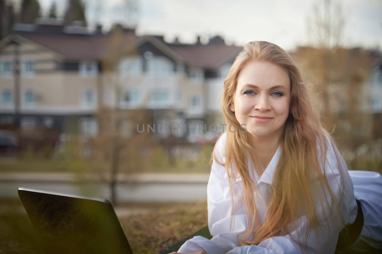 Beautiful blonde girl working by laptop in gazebo on an autumn, summer or spring day. Businesswoman, student, freelancer or manager works outdoors in natural landscape in village, city or town