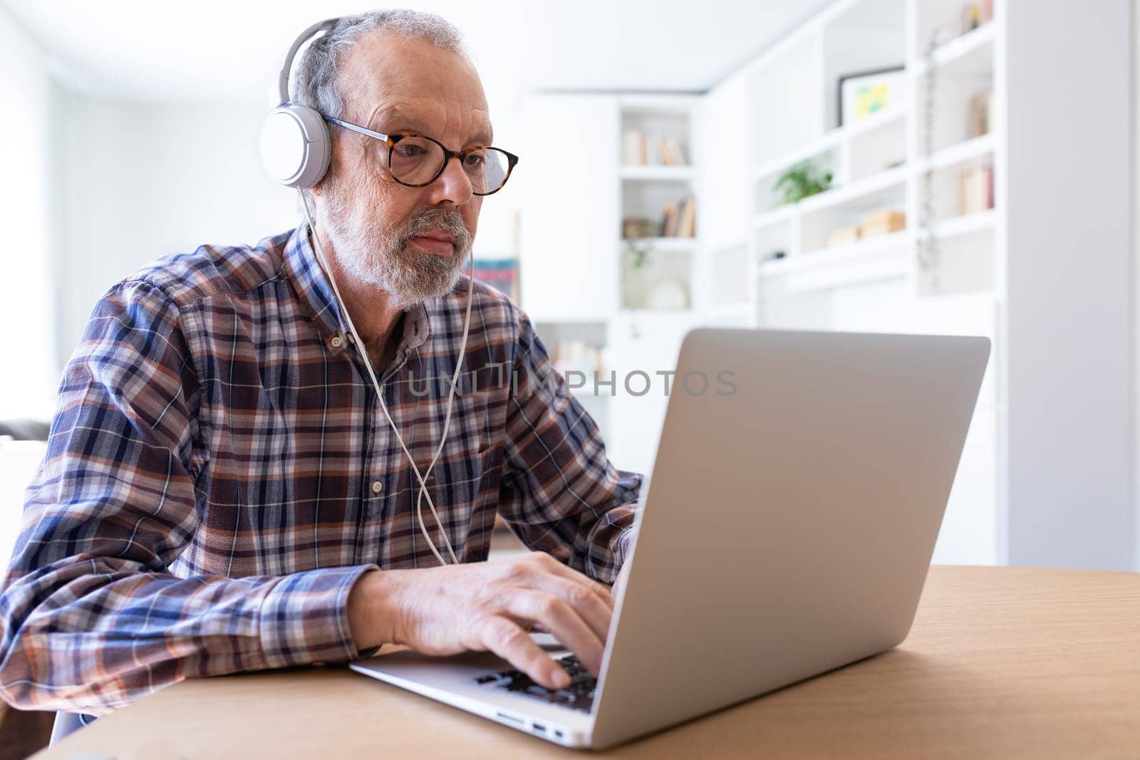 Senior man working with laptop and using headphones at home dining room. Lifestyle and technology concepts.