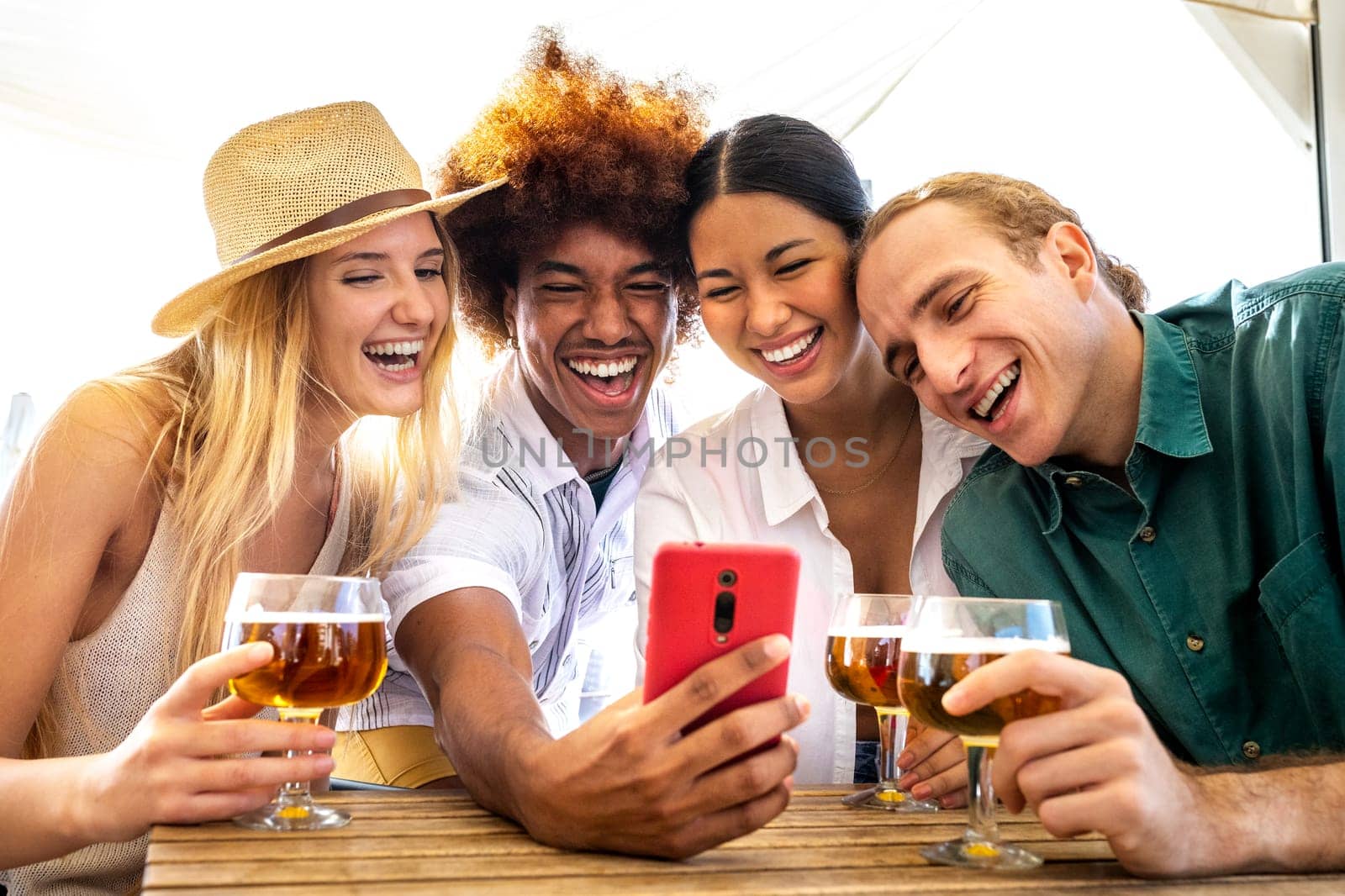 Group of happy multiracial friends looking at mobile phone in a beach bar while drinking beer. Friends on a video call using phone. Taking selfie. Technology and leisure activity.