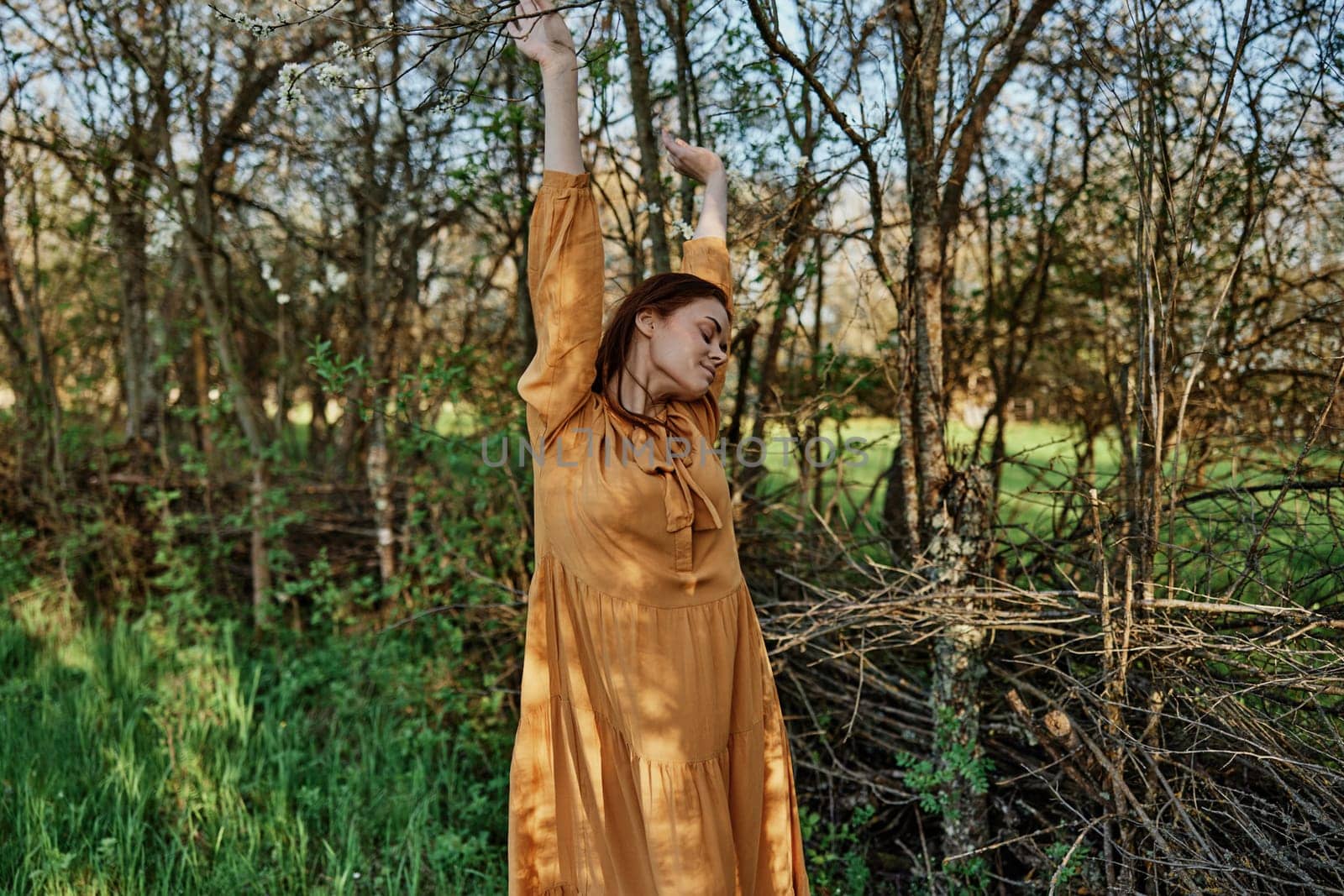 a beautiful, slender woman with long hair walks in the shade near the trees, dressed in a long orange dress, enjoying the weather and the weekend, joyfully raising her hands up. High quality photo